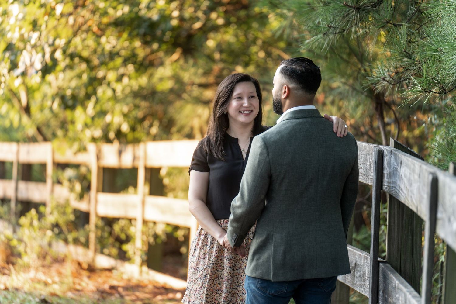 A couple stands together in an outdoor setting with autumn foliage and sunlight filtering through trees.