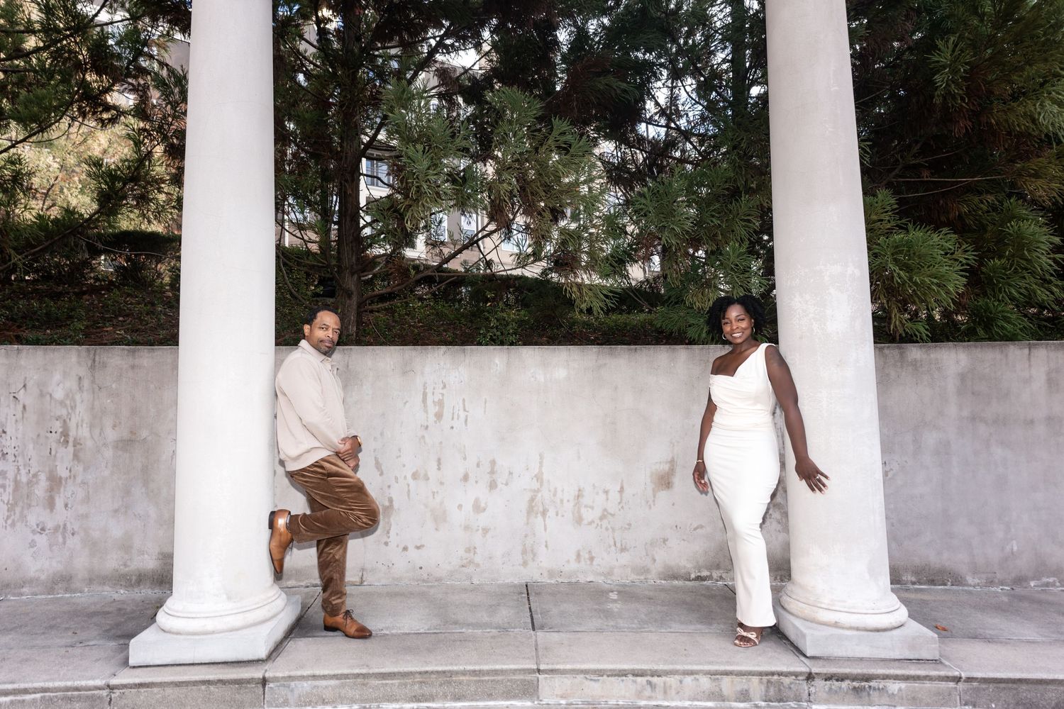 Two people pose elegantly against white classical columns in an outdoor garden setting.