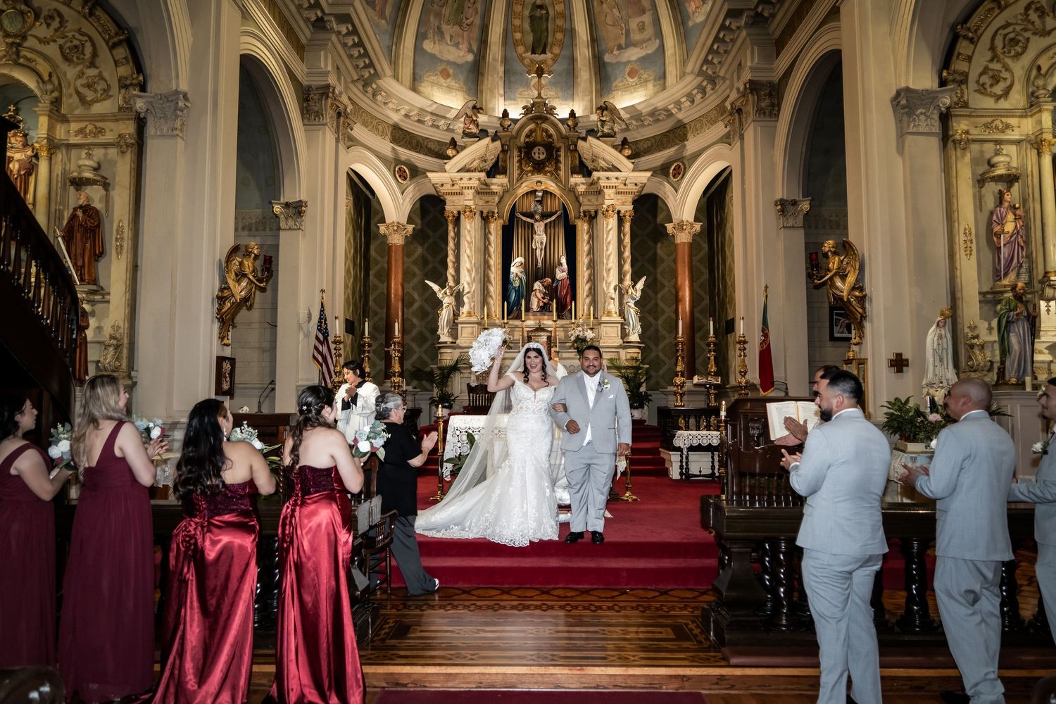 Wedding ceremony taking place in an ornate Catholic church with burgundy-clad bridesmaids and groomsmen in light blue suits.