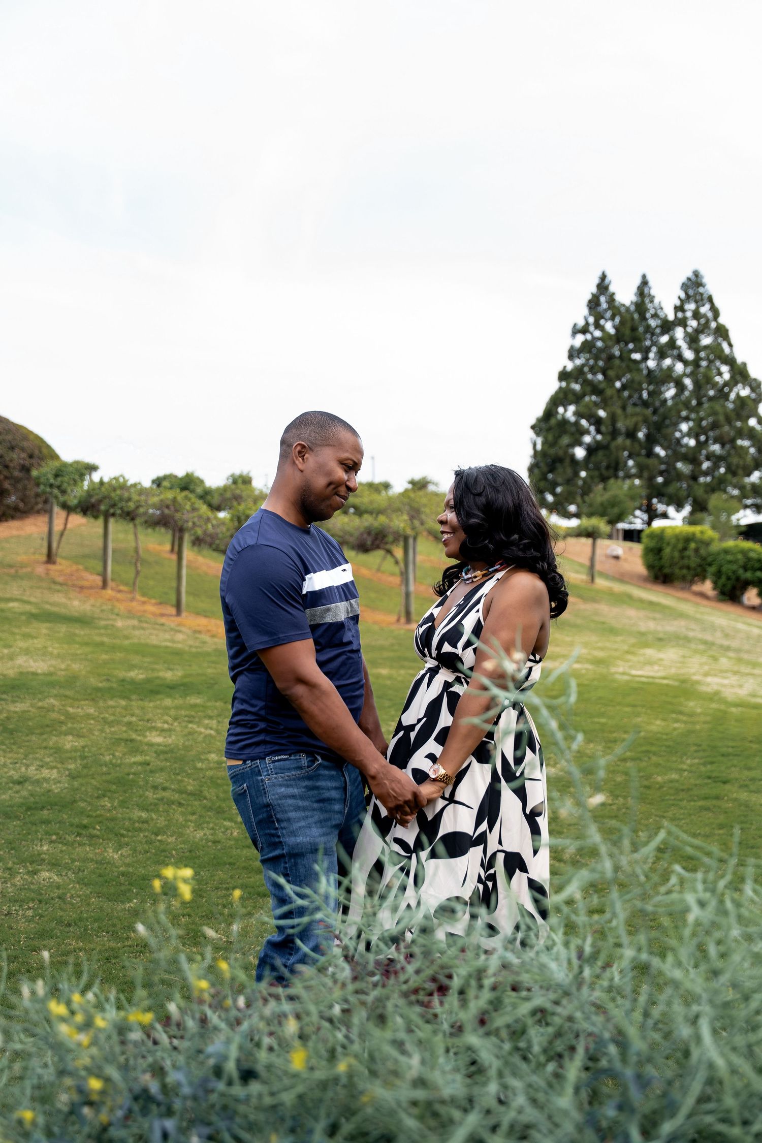 A couple shares a romantic moment together on a grassy hillside with evergreen trees in the background.