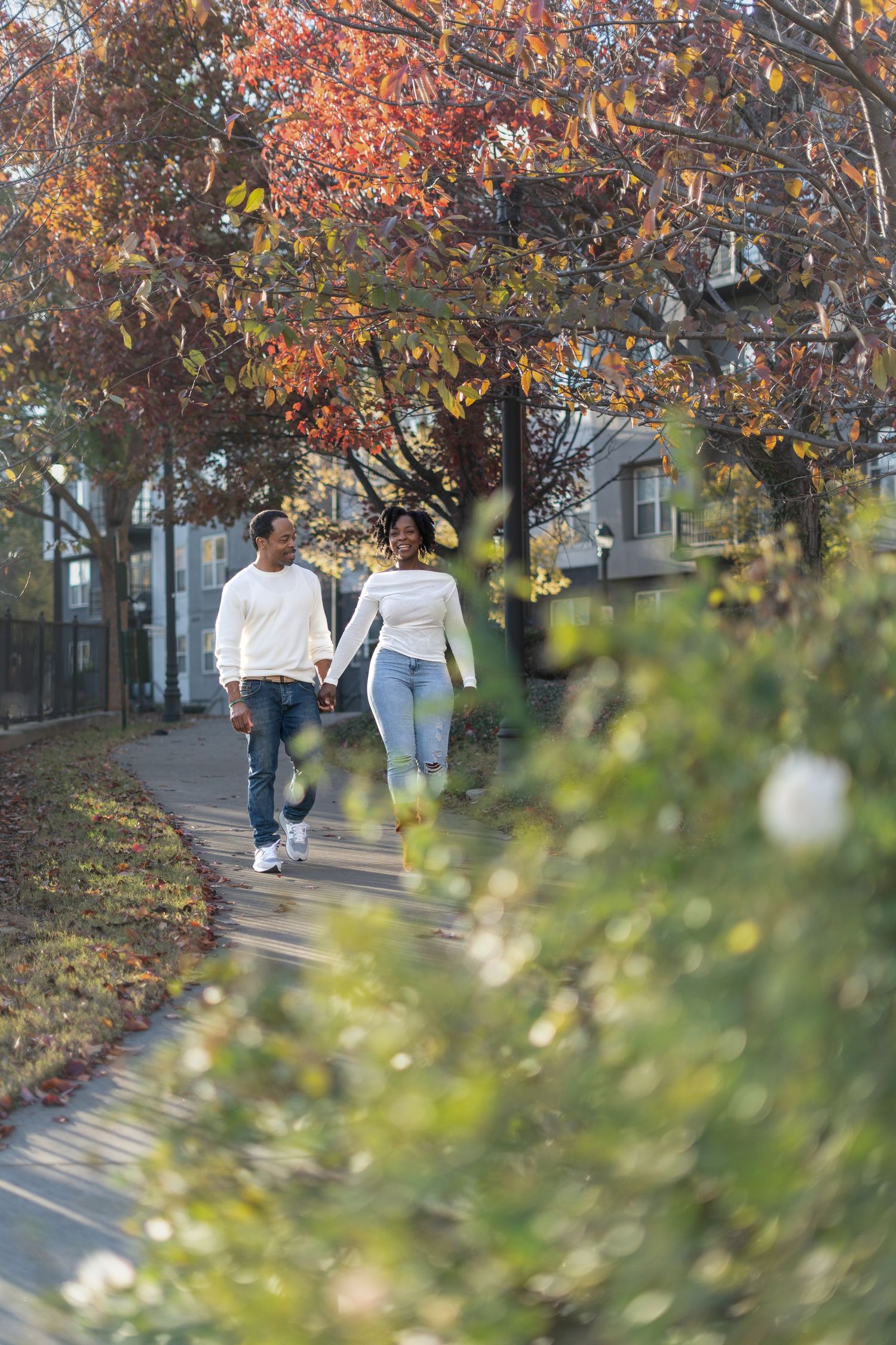A couple walks together along a sidewalk under autumn trees with orange and red foliage on a fall day.
