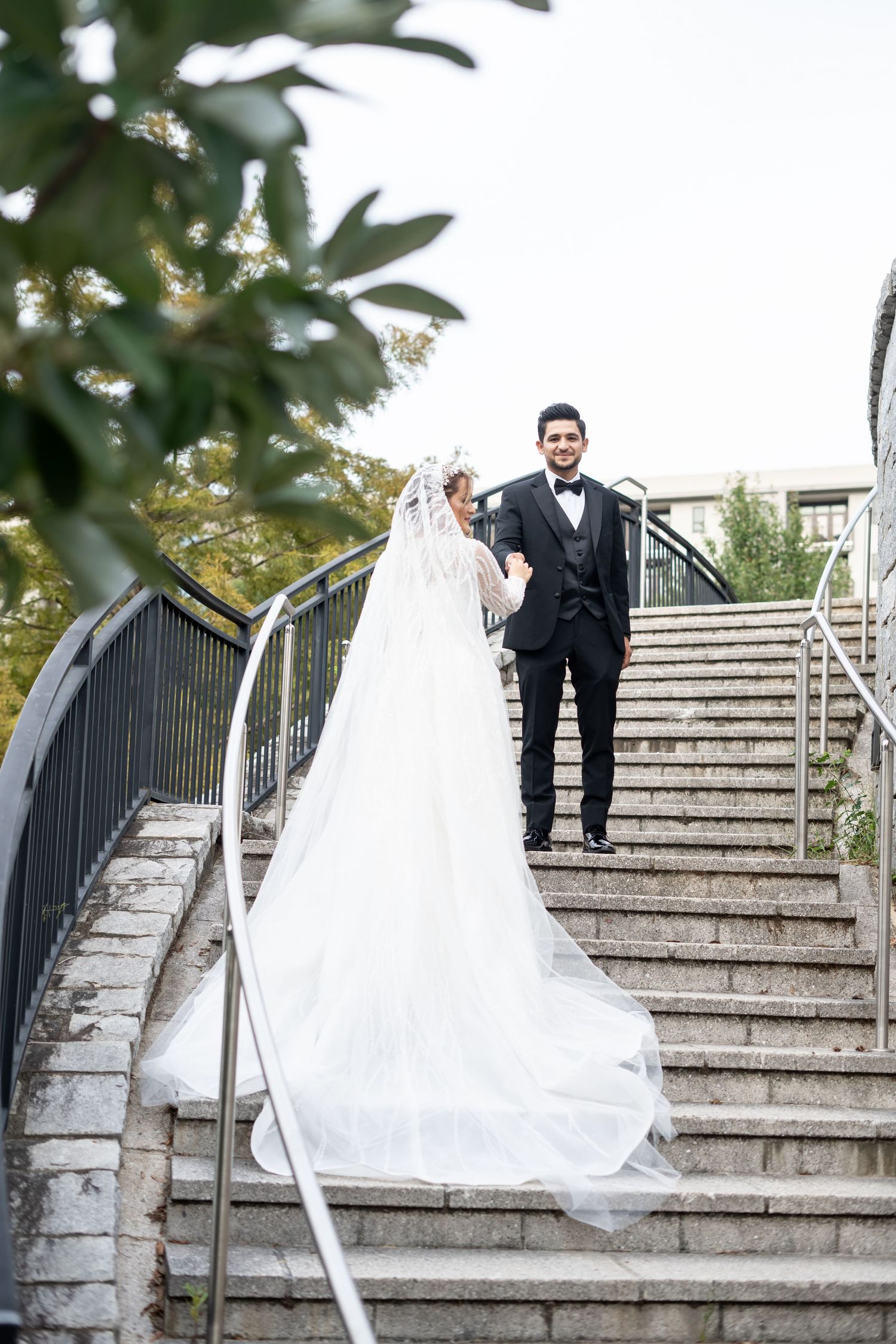 A wedding couple poses on elegant stone steps with white railings and greenery in the background.