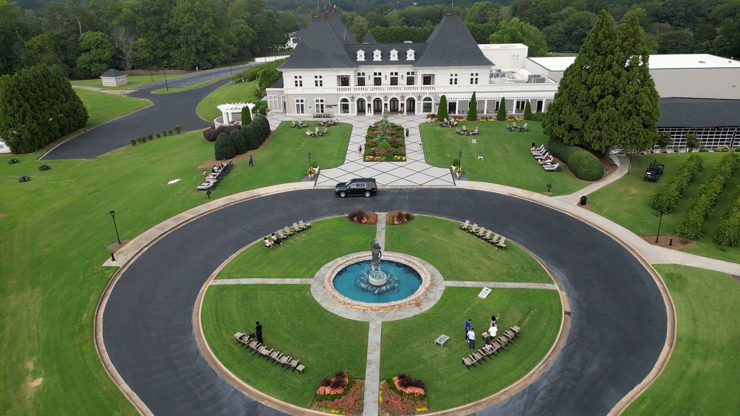 Aerial view of an elegant estate with a circular fountain driveway, manicured lawns, and a grand white mansion.