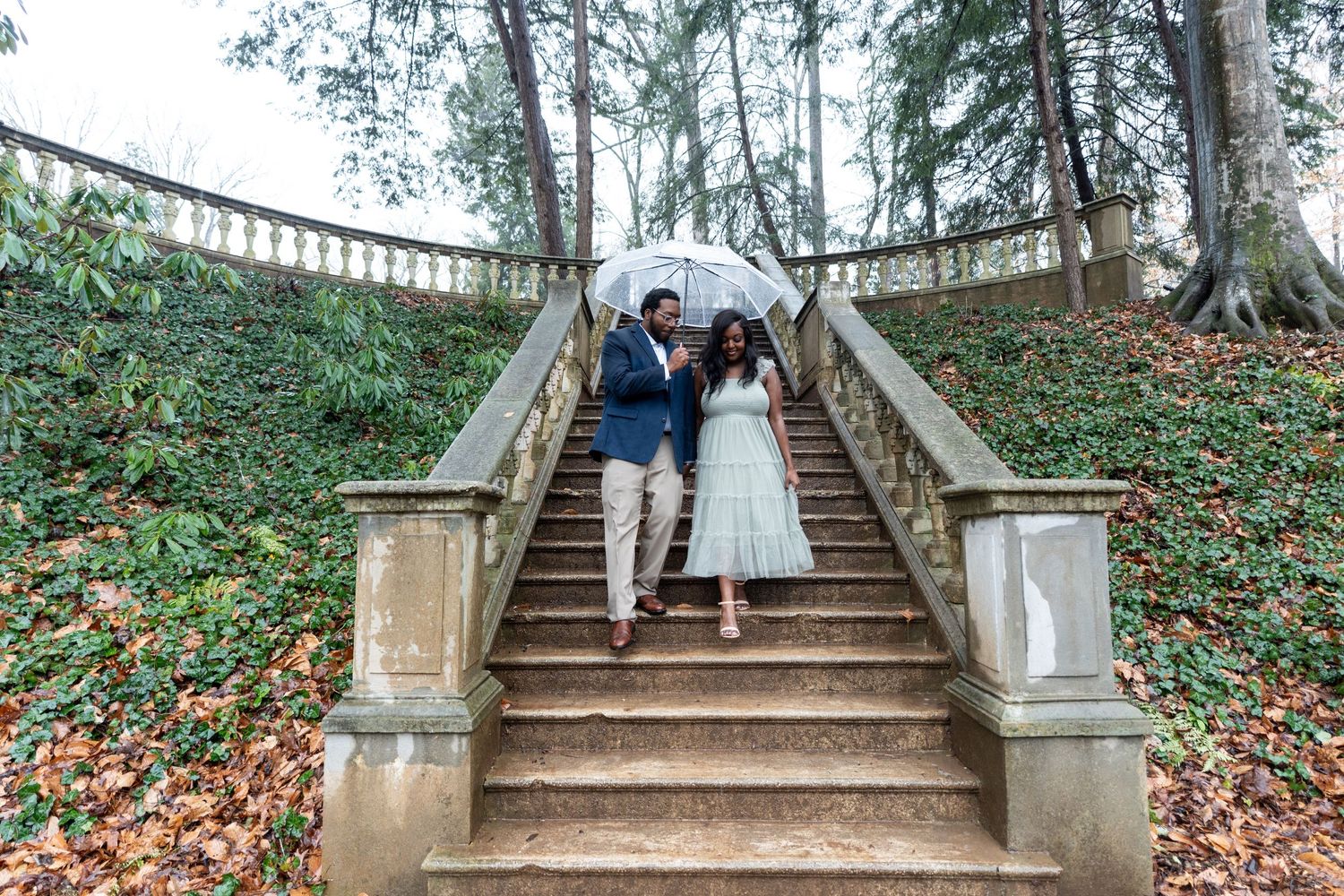 A couple in wedding attire poses on an elegant stone staircase surrounded by autumn leaves in a garden setting.