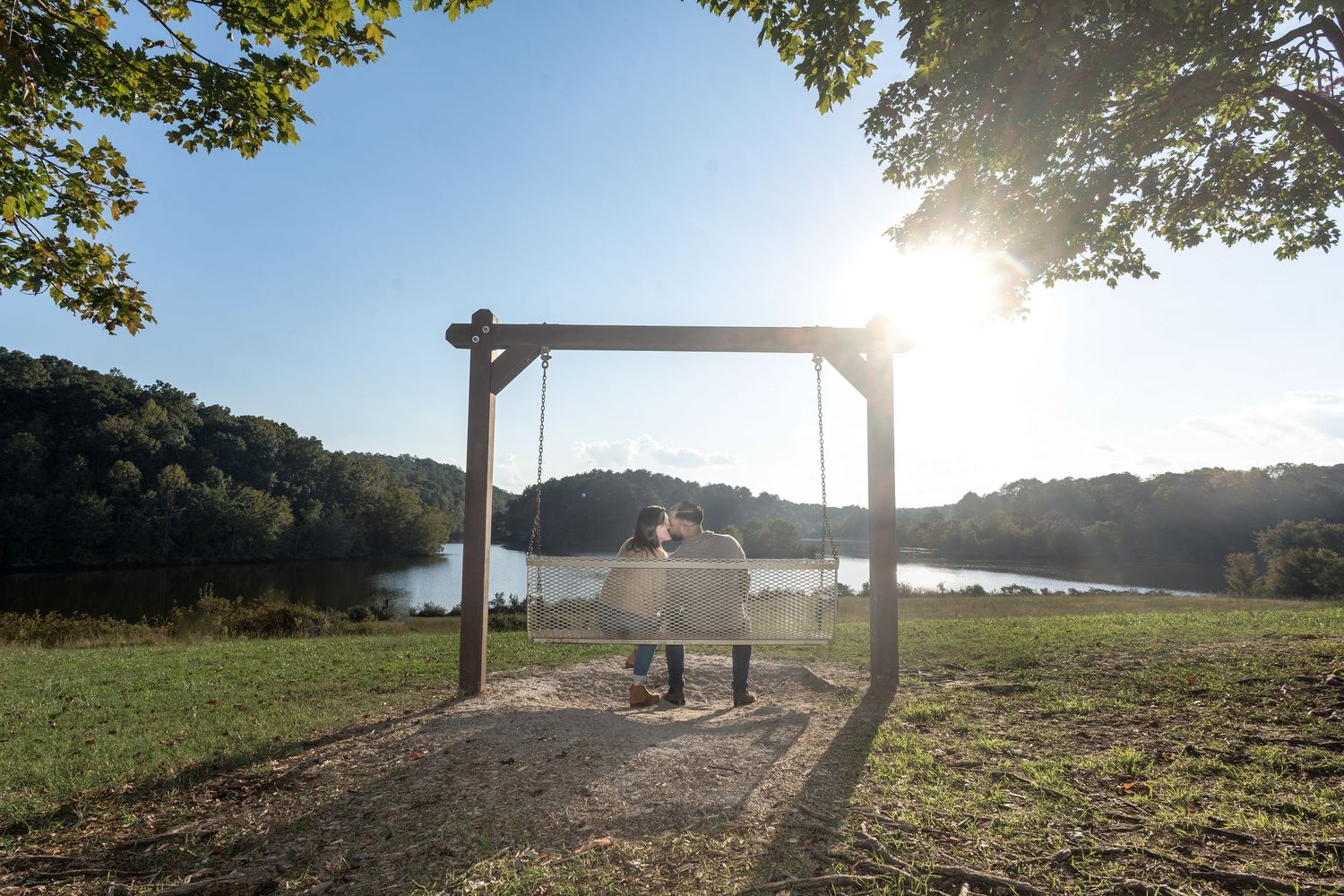 Scenic panoramic view of a misty lakeside park bench at sunrise with golden sunlight filtering through trees.