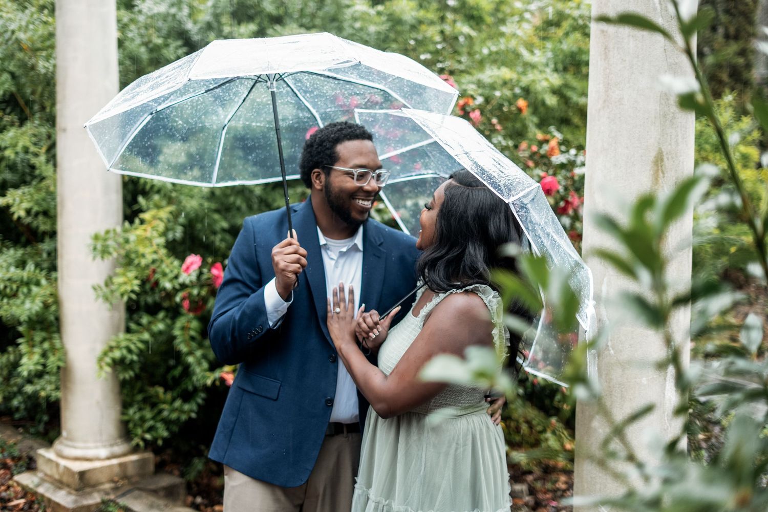 A couple shares an intimate moment under a clear umbrella in a lush garden setting with white pillars.
