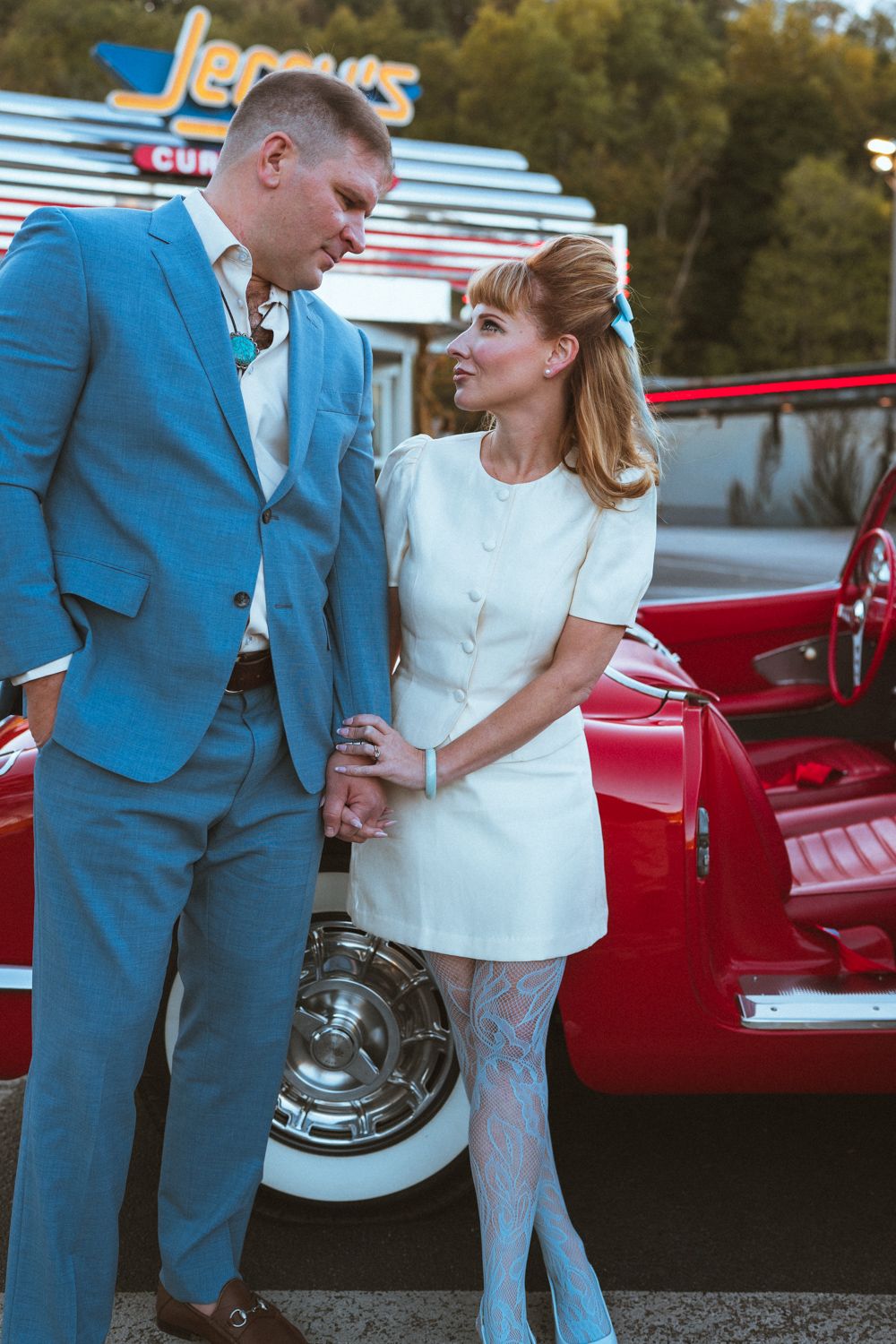 A couple in retro clothing poses together near a vintage red car in a romantic photoshoot series.