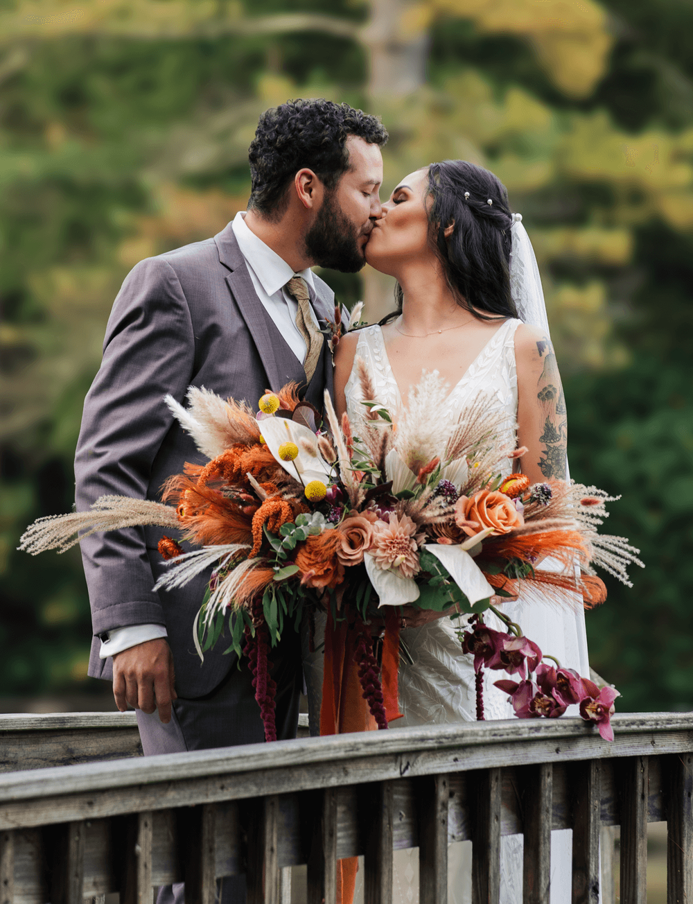 Bride and groom share a romantic kiss on a wooden bridge, bride holding a lush autumn bouquet with orange and rust blooms.