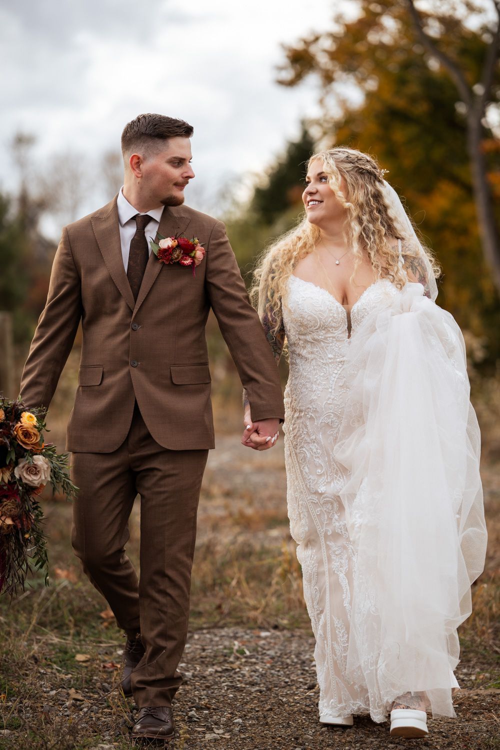 Bride and groom walk hand-in-hand outdoors in autumn, she in lace gown, he in brown suit with floral boutonniere.