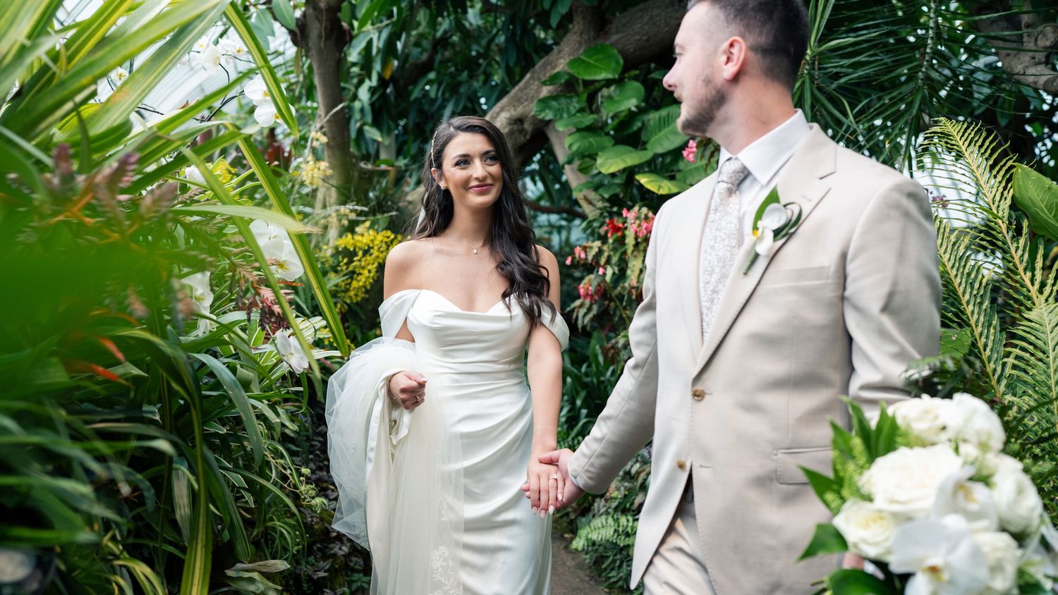 A bride in a white gown and groom in a beige suit hold hands smiling in a lush tropical garden setting.