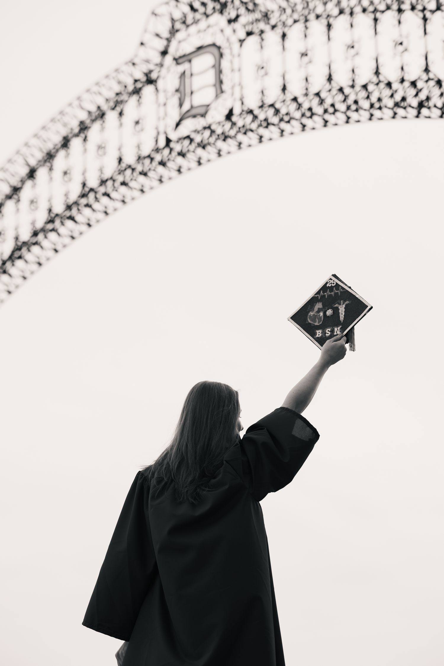 Black and white photo of a graduate in cap and gown raising diploma toward decorative arch outdoors.