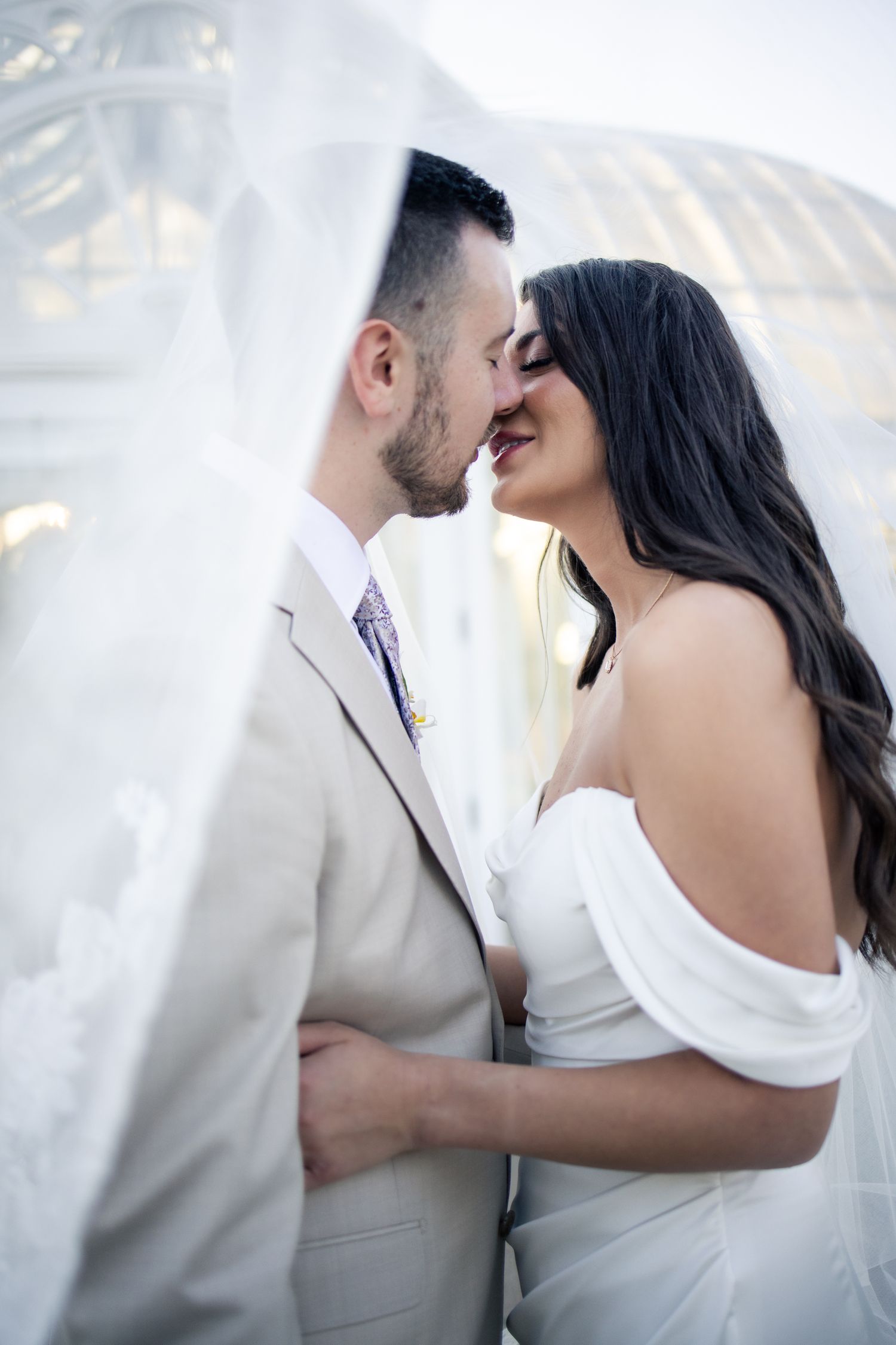 Bride and groom share a romantic almost-kiss under a flowing white veil on their wedding day.