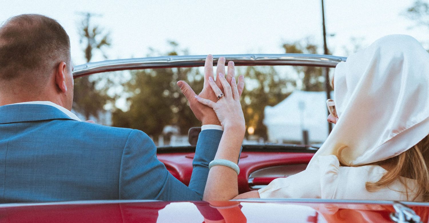 Newlywed couple sitting in a red convertible car, holding hands and showing wedding rings on their special day.