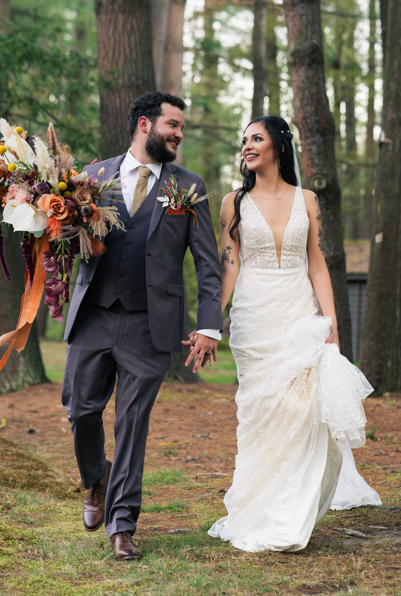 Smiling bride and groom walk hand-in-hand through pine forest, bride in lace gown, groom in gray suit with bouquet.