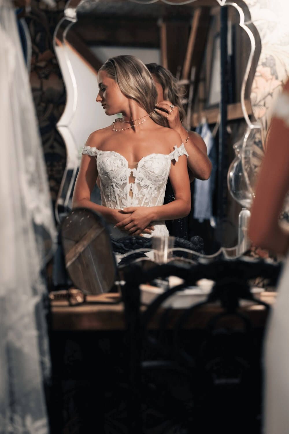 Bride in white lace off-shoulder corset gown adjusting hair jewelry reflected in ornate mirror.