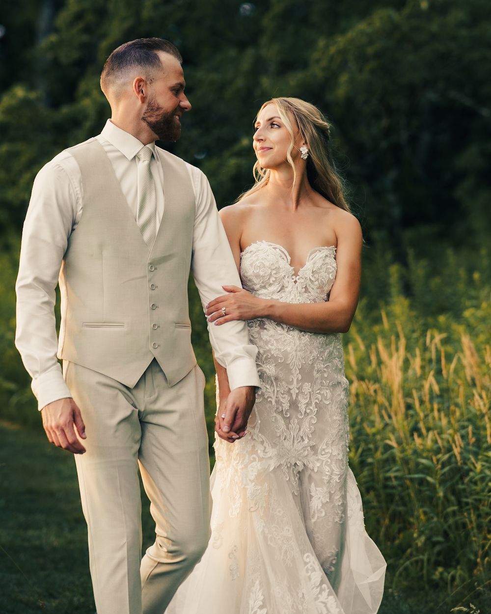 A couple in wedding attire walks hand-in-hand outdoors, the groom in a beige suit and bride in a lace gown.