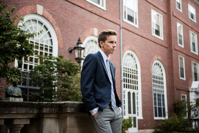 A young professional in a navy blazer and gray slacks walks past an elegant brick building with arched windows.