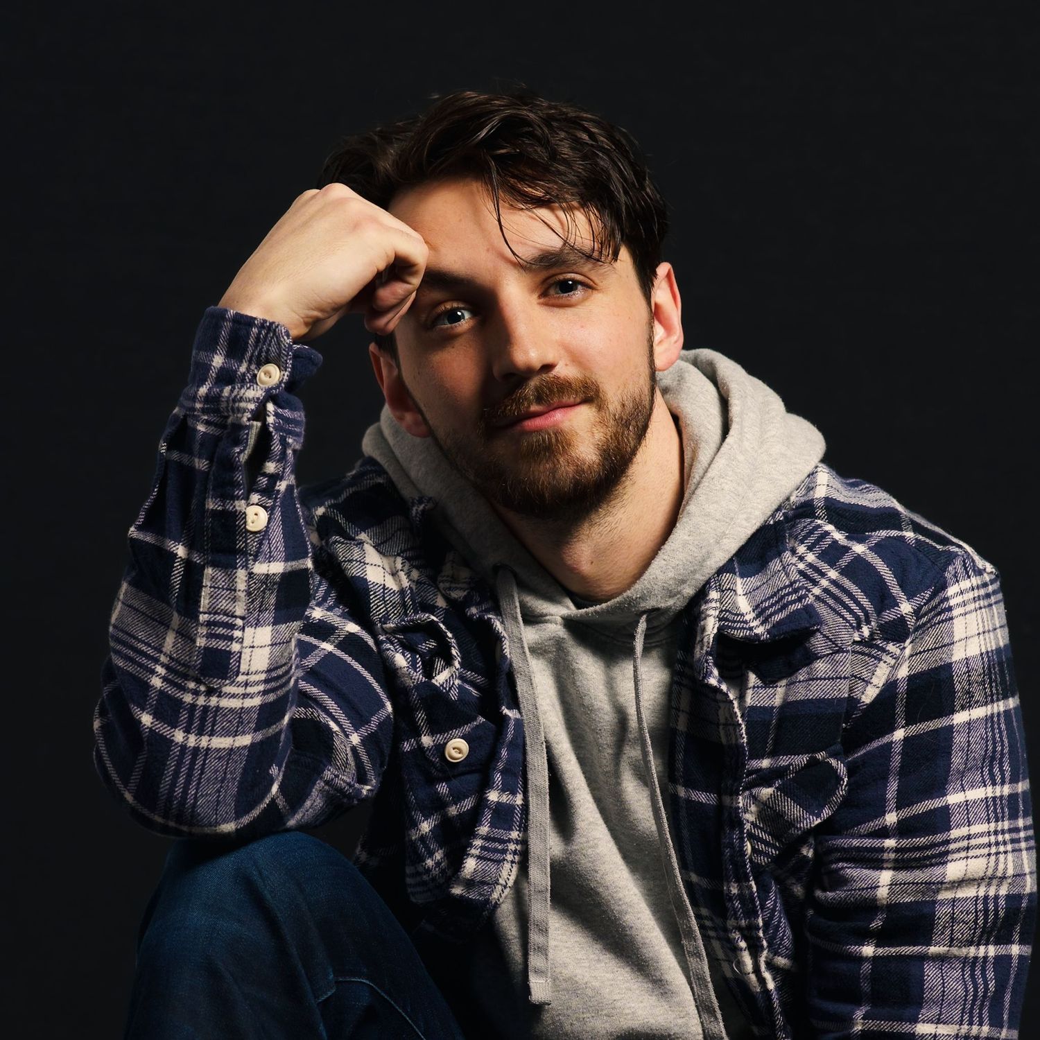 Young man with beard in flannel shirt and hoodie posing against dark background with hand on head.