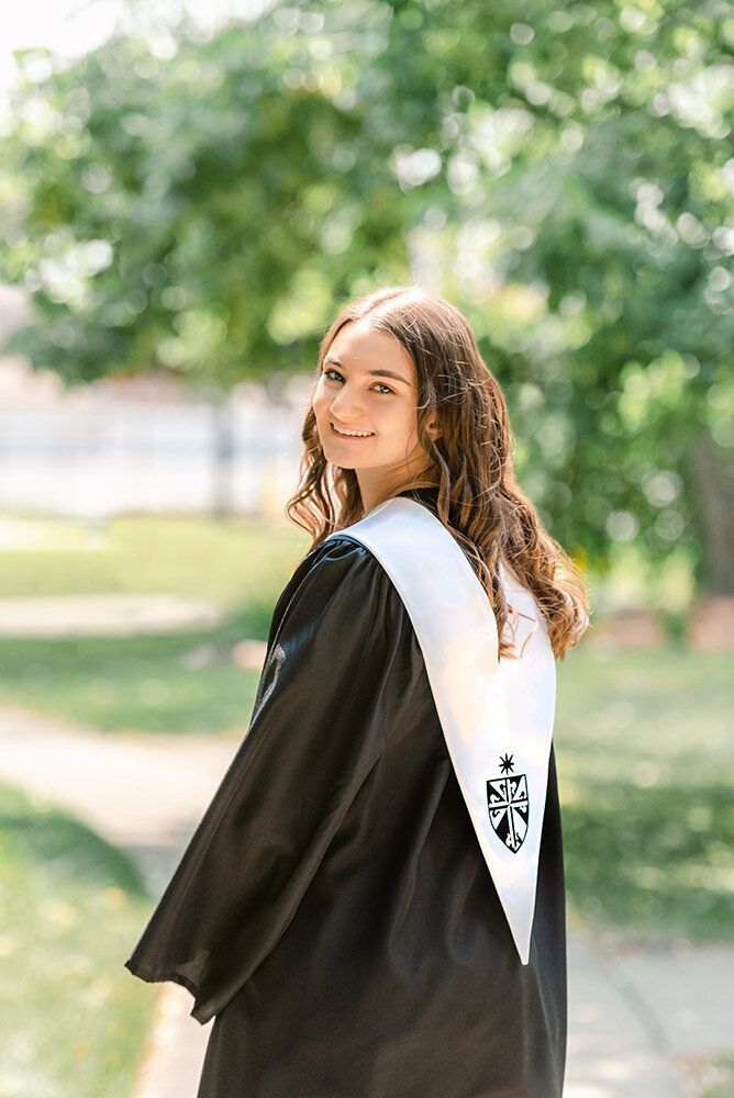 Graduate in black cap and gown with white sash poses outdoors on sunny day with trees in background.