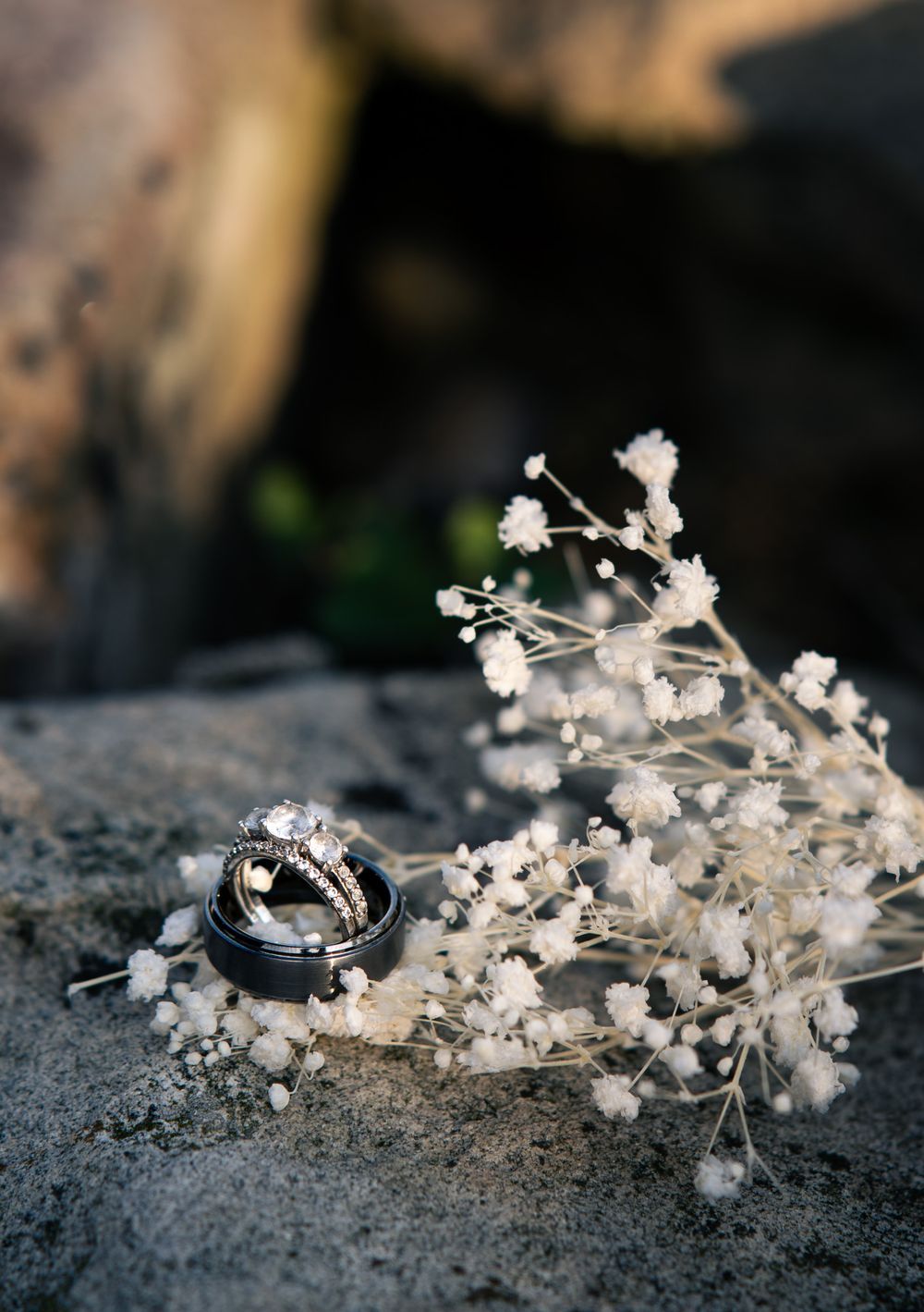 Wedding rings and diamond engagement ring nestled in baby's breath flowers on a rocky surface.