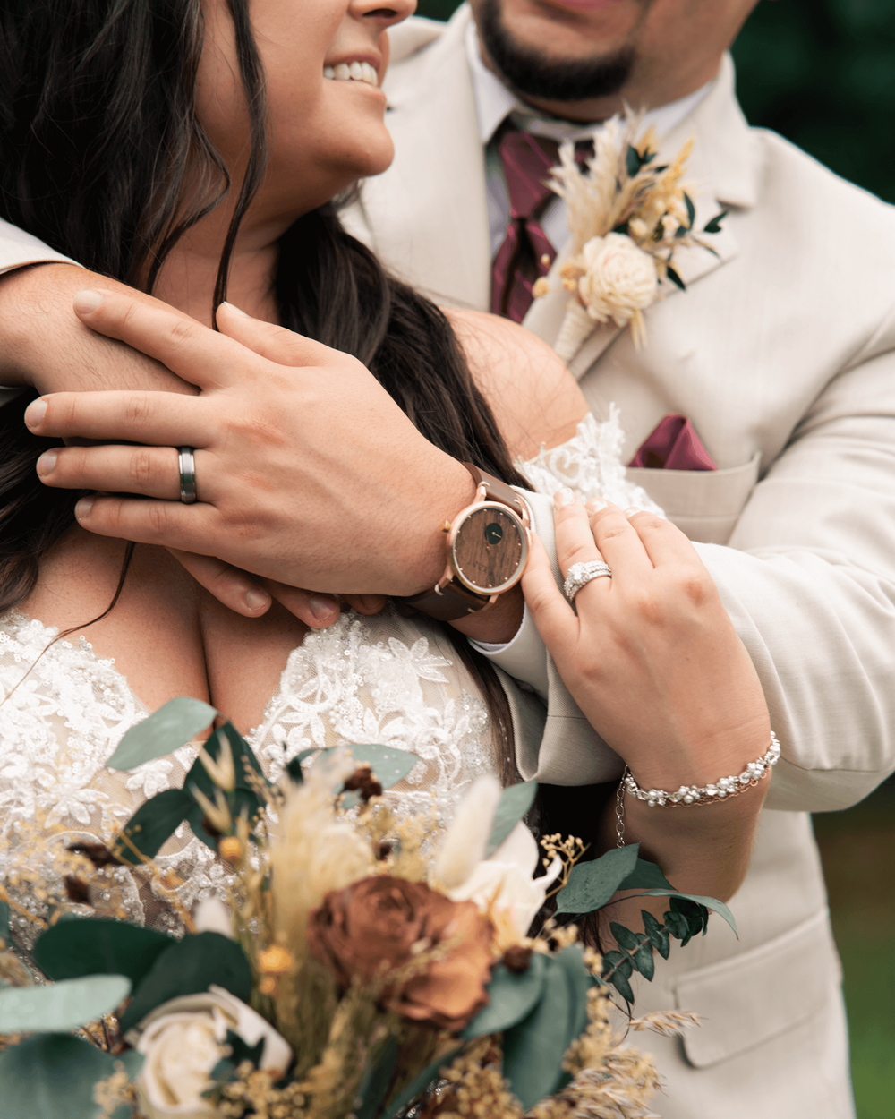 Bride and groom embrace, showcasing wedding rings, a wood-face watch, and a floral bouquet with eucalyptus at their wedding.