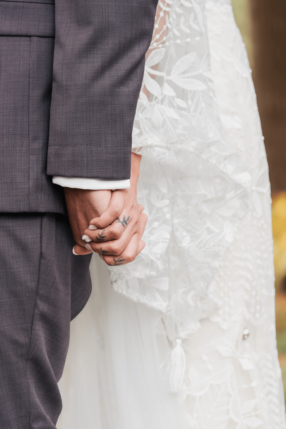 Close-up of bride and groom holding hands, showcasing lace wedding dress and gray suit with tattooed fingers.