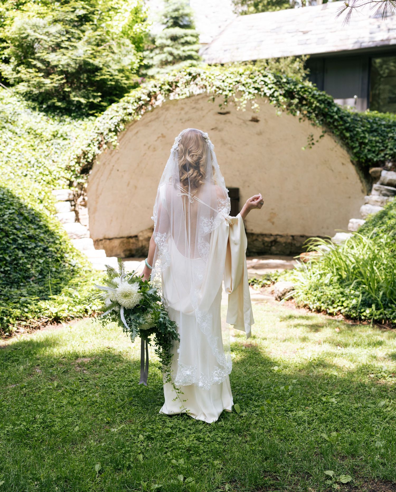 Bride in white gown and flowing veil stands in lush garden holding bouquet near stone arch structure.