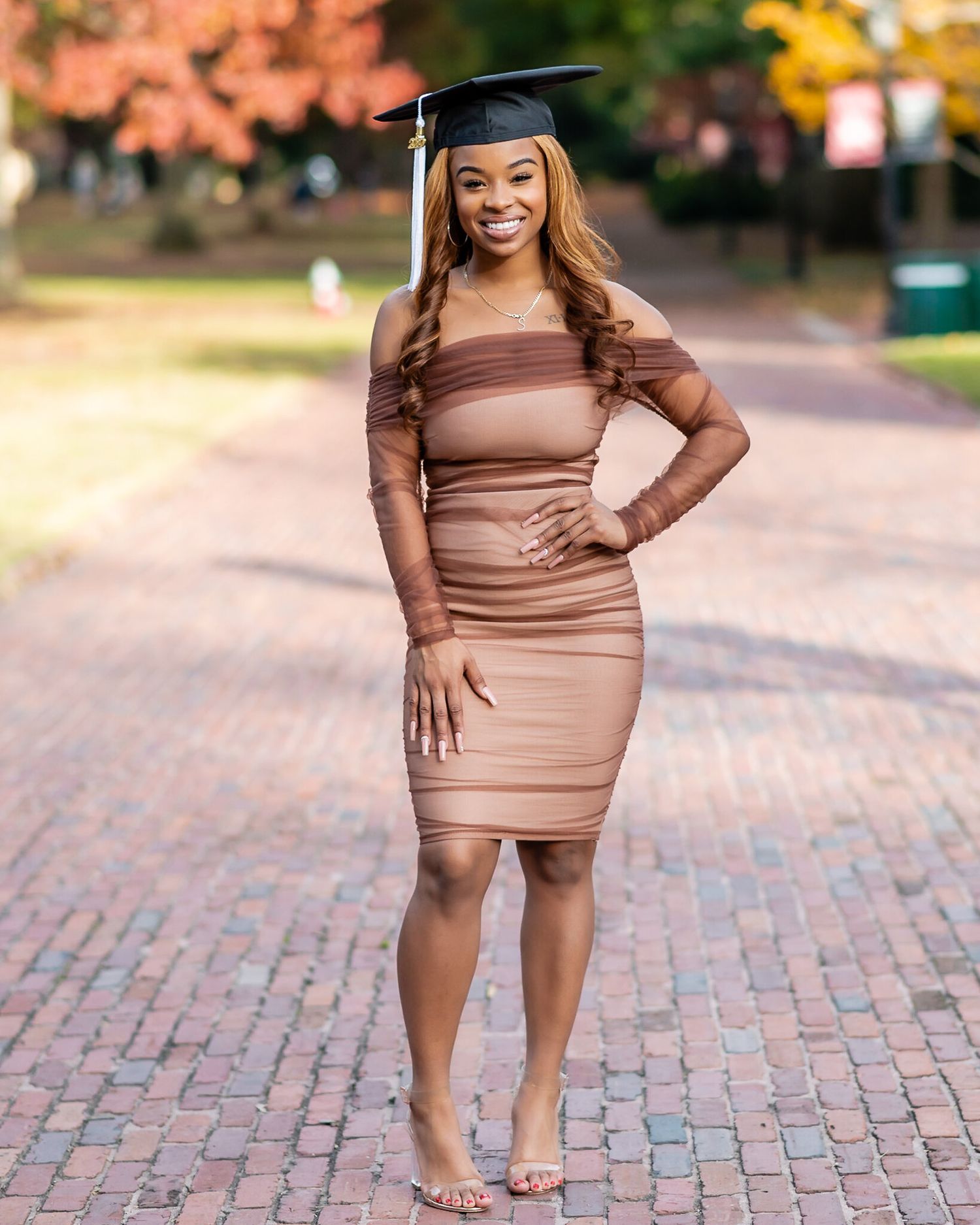 Graduate in brown dress and mortarboard cap stands on brick pathway with autumn trees in background.
