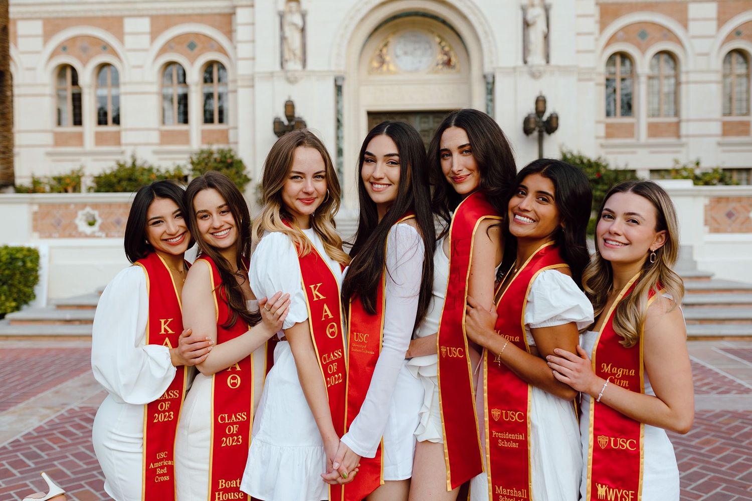 Seven graduates in white dresses and red graduation sashes pose together on steps of ornate building.
