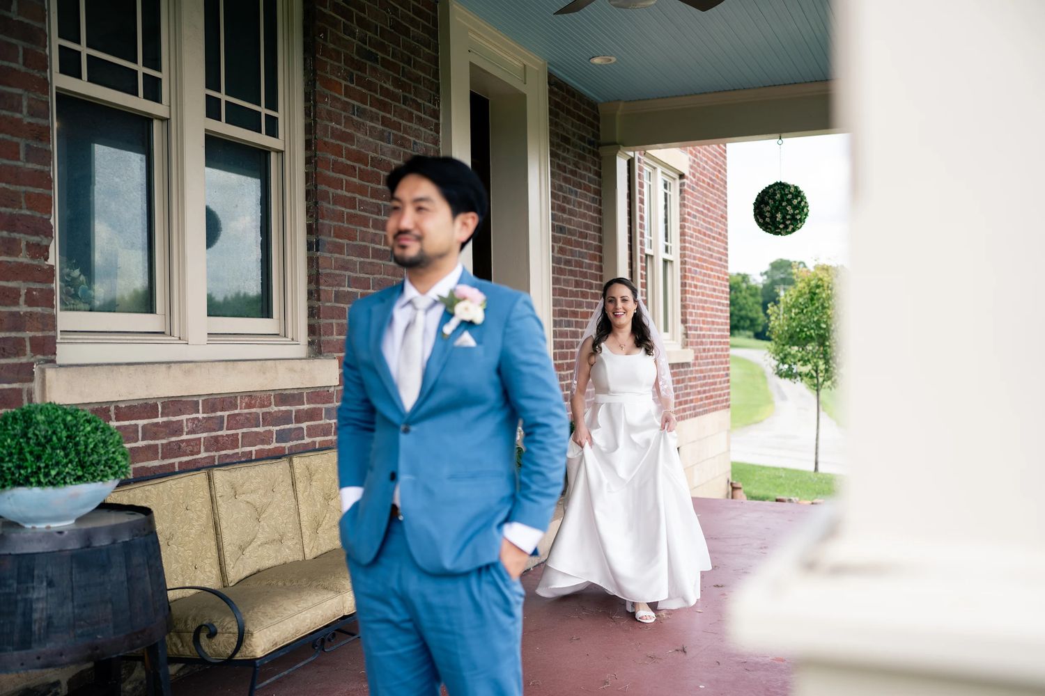 Groom in blue suit waits on brick porch as bride in white gown approaches during wedding first look.