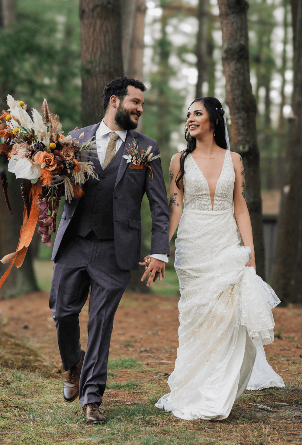 A smiling bride and groom walk hand-in-hand through a forest, she in a lace gown, he in a gray suit with bouquet.