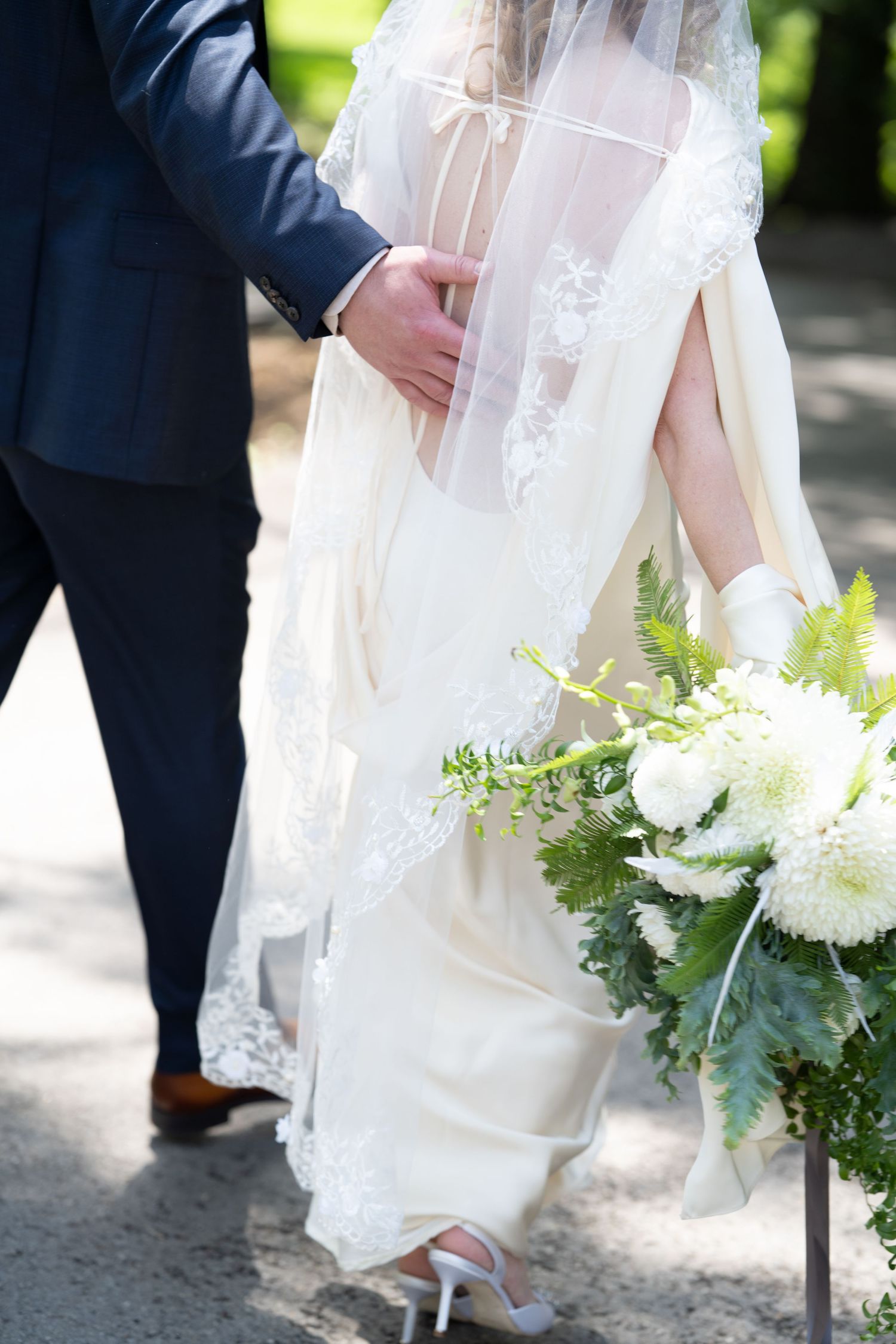 Bride in elegant lace veil and ivory gown walks with groom in navy suit, holding white floral bouquet outdoors.
