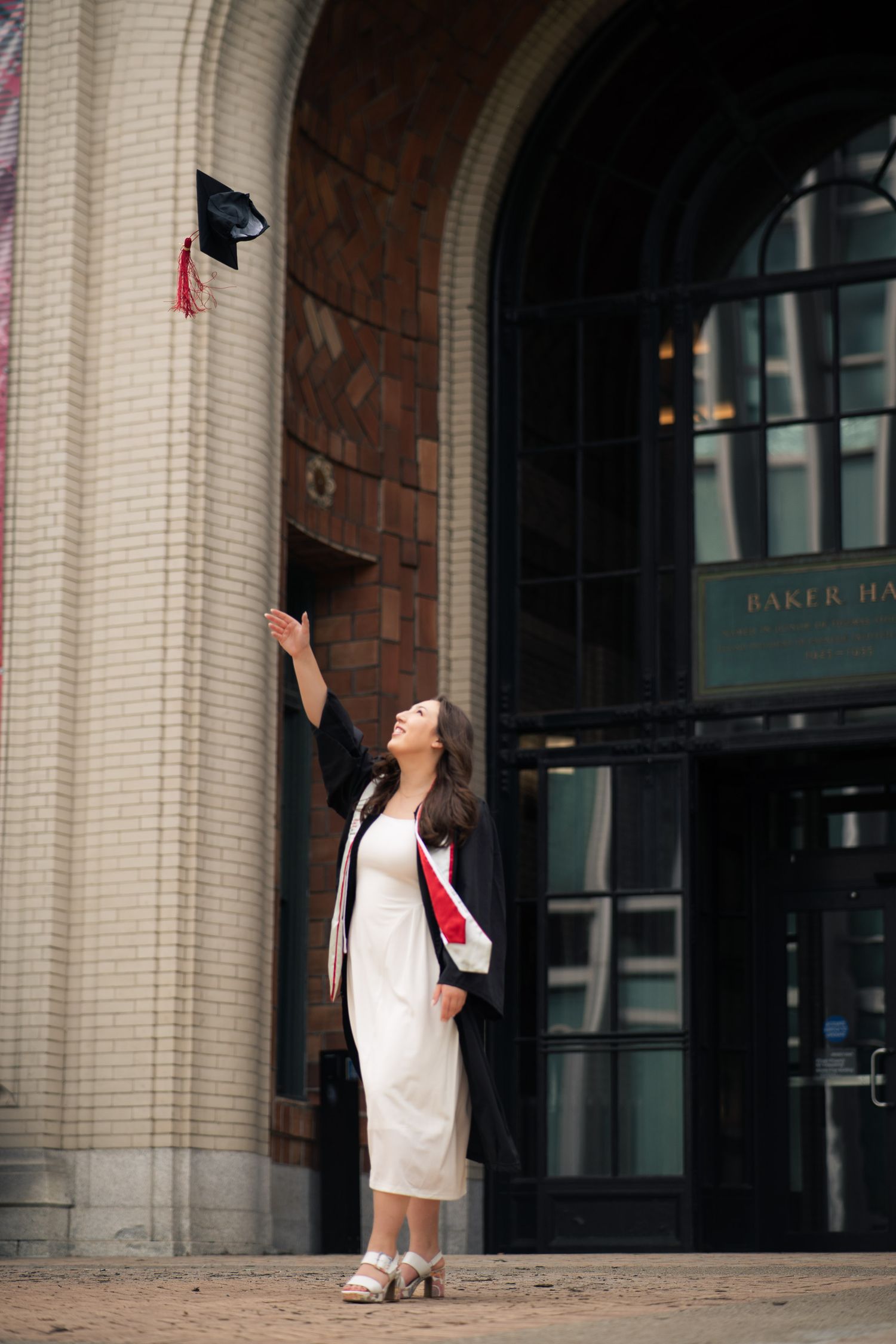 Graduate in white dress and black gown tosses cap in front of Baker Hall building entrance.