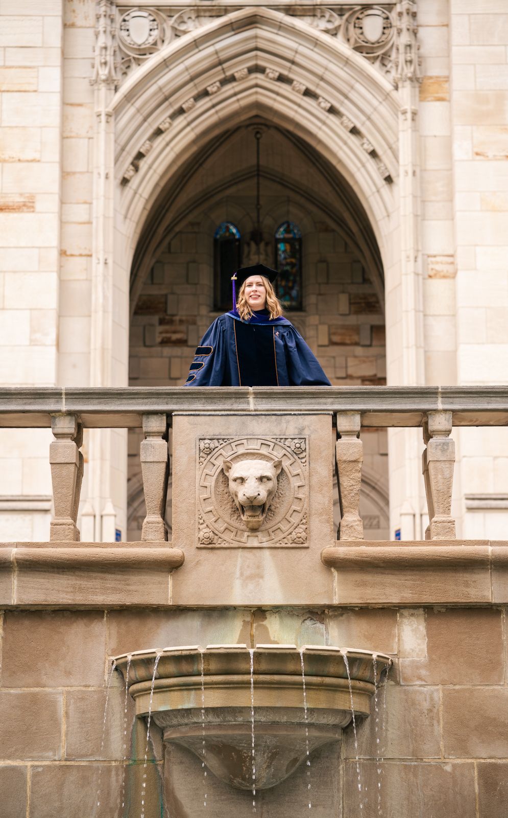 Graduate in academic regalia stands on ornate stone balcony with lion fountain at a Gothic university building.
