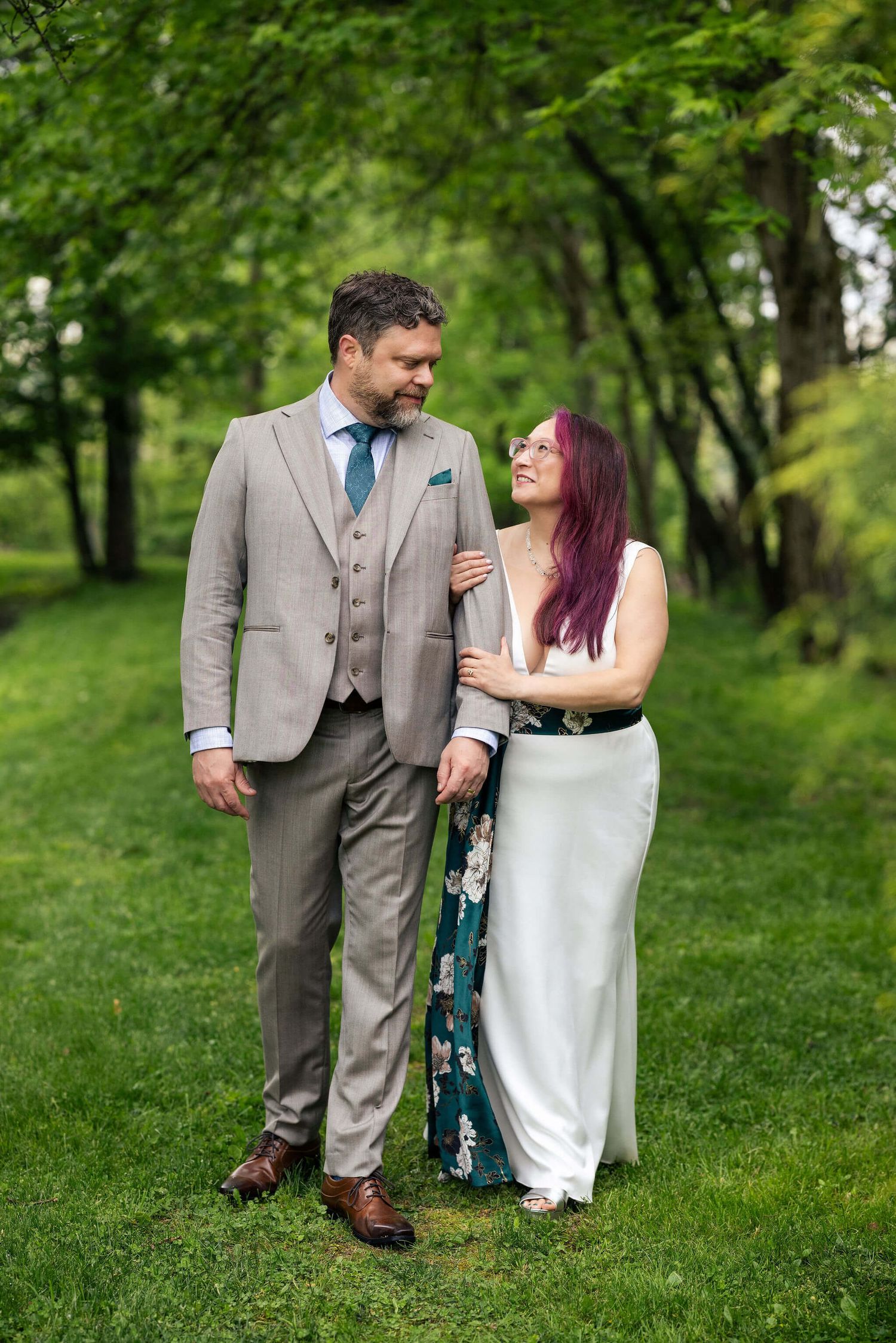 A couple in wedding attire smiles at each other while walking through a lush green park setting.
