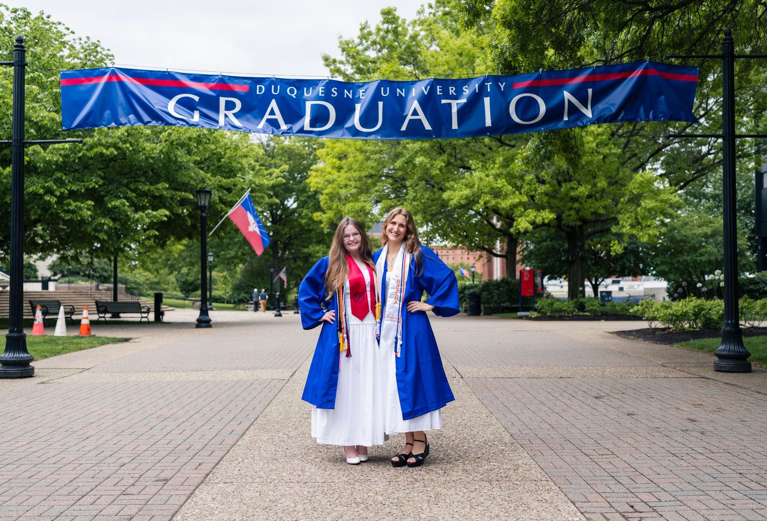 Two graduates in blue robes pose under a Duquesne University Graduation banner outdoors.