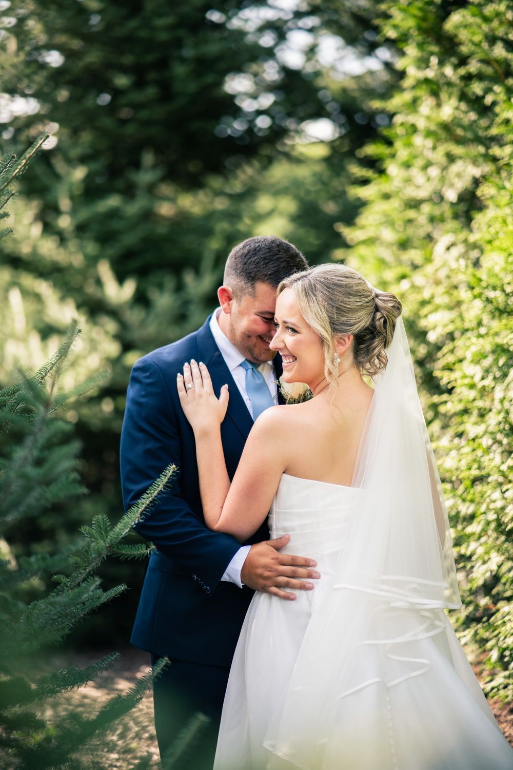 A romantic wedding couple embraces outdoors in a lush garden setting with flowing veil and navy suit.