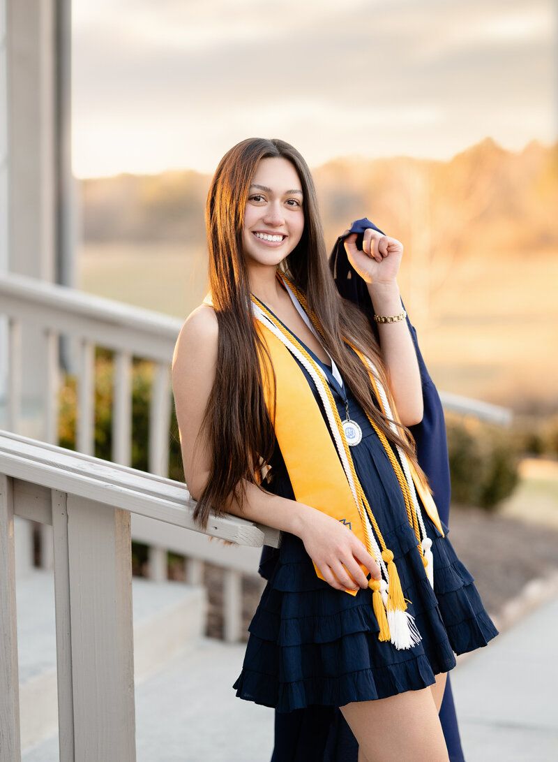 Graduate in navy dress and gold sash stands on porch railing during golden sunset while celebrating graduation.