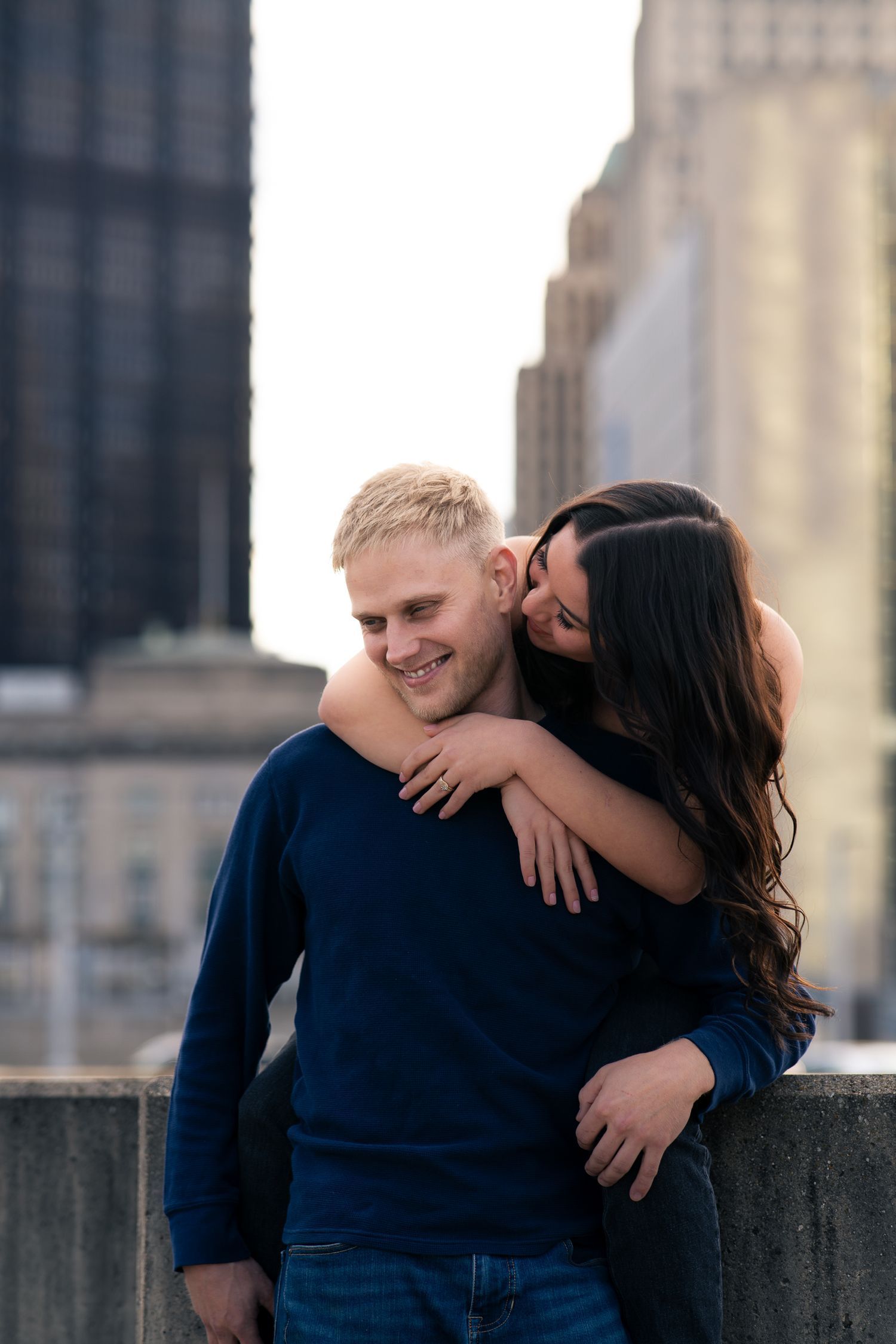 A couple embraces on a rooftop with city buildings in the background, woman kissing mans cheek while he smiles.