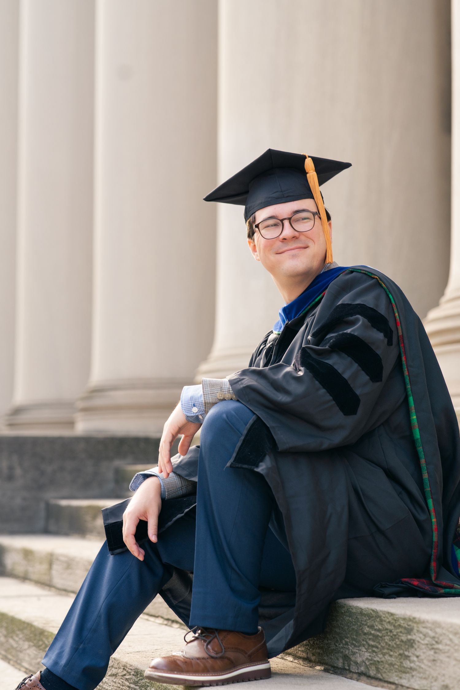 A smiling graduate in black cap and gown with gold tassel sits on stone steps beside tall columns.