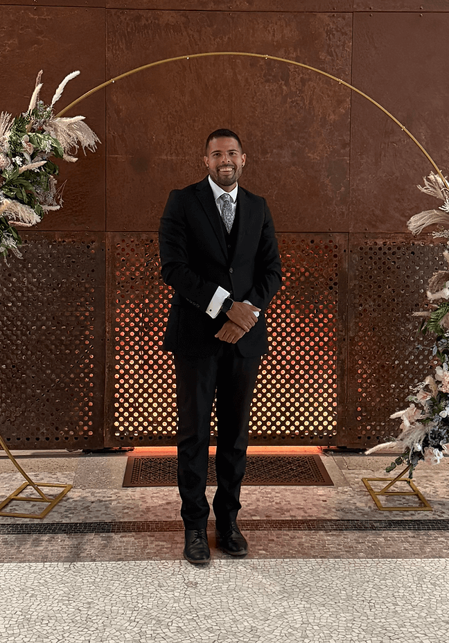 A person in a black suit sits on a rustic brown wall with lattice and floral decorations in the background.