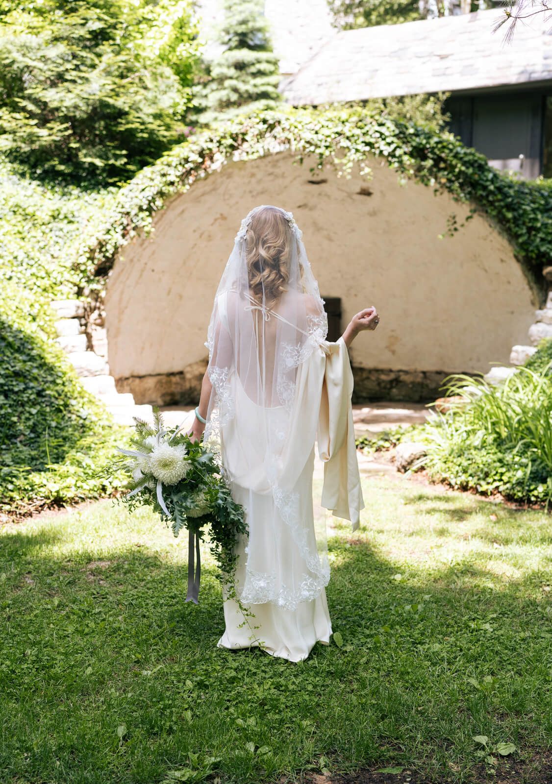 Bride in white gown and lace veil holds bouquet, standing in lush garden with ivy-covered stone structure.