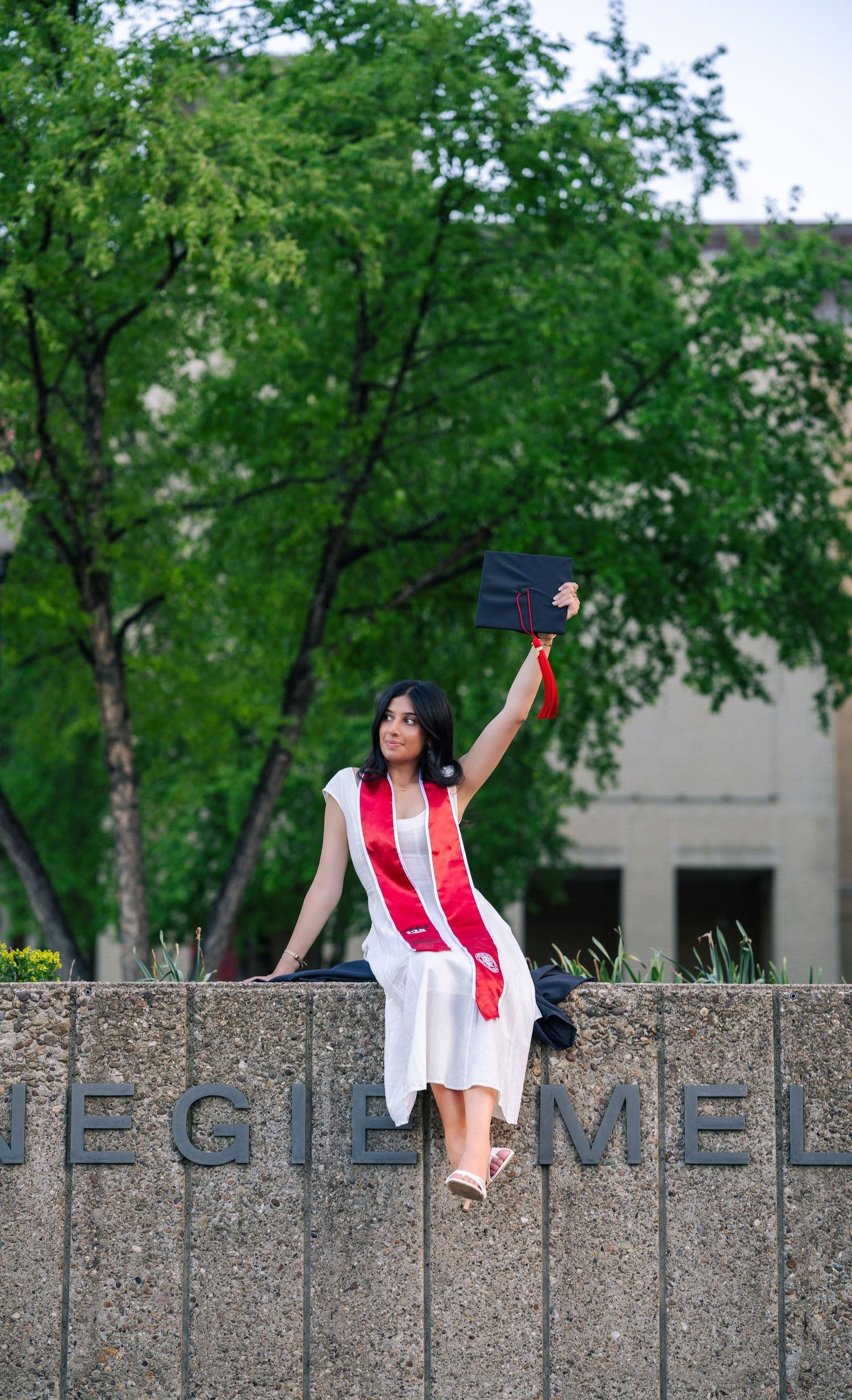 A graduate in white dress and red sash raises her cap triumphantly while sitting on a Carnegie Mellon University stone wall.