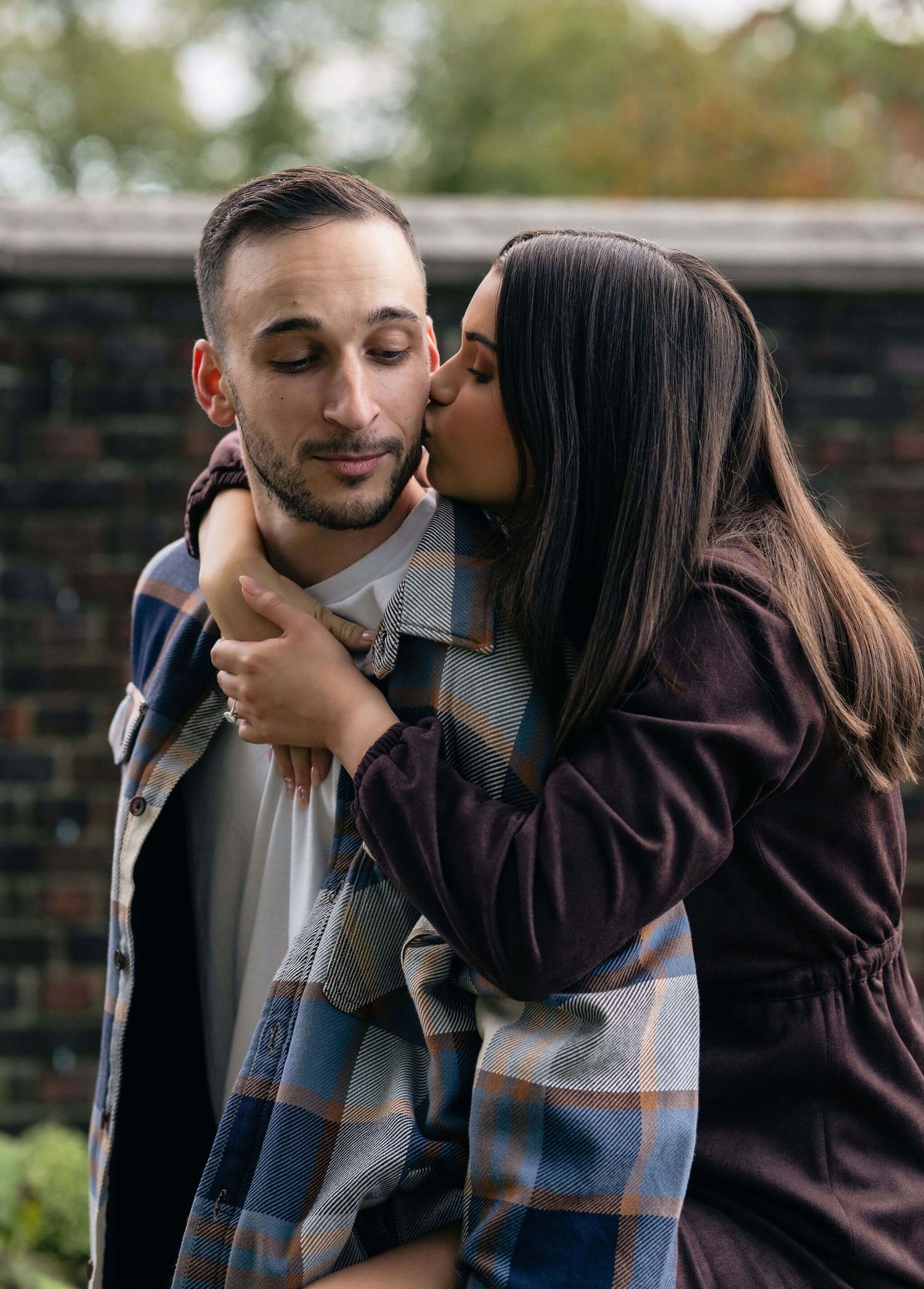 A woman with long dark hair kisses a man on the cheek while embracing him outdoors near a brick wall.