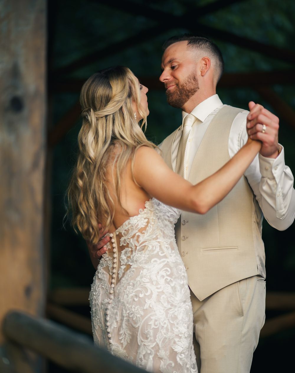 A bride and groom share their first dance, she in lace gown, he in tan suit, gazing lovingly at each other.
