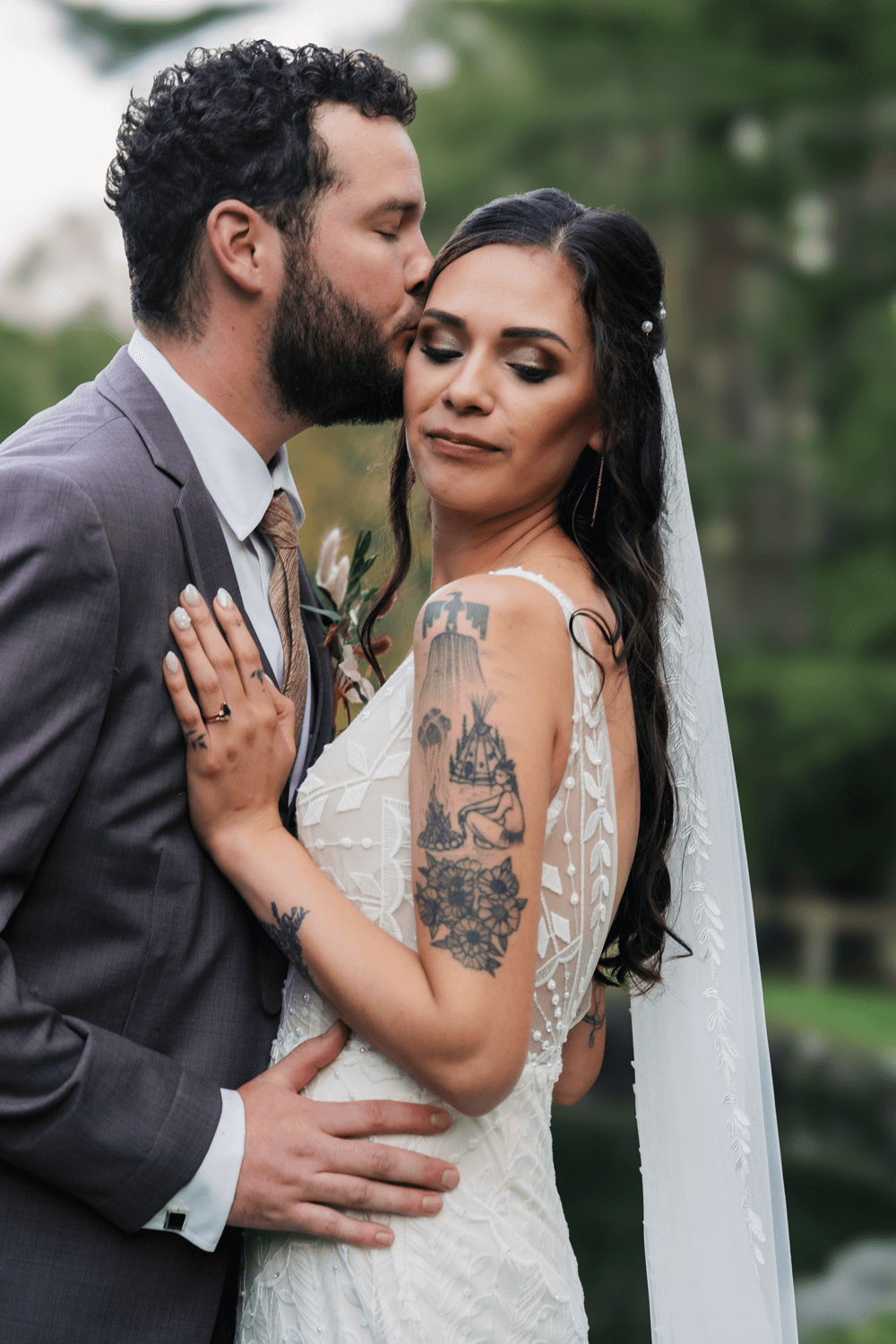 A groom in a gray suit kisses his tattooed bride in a white lace gown and veil during an outdoor wedding.