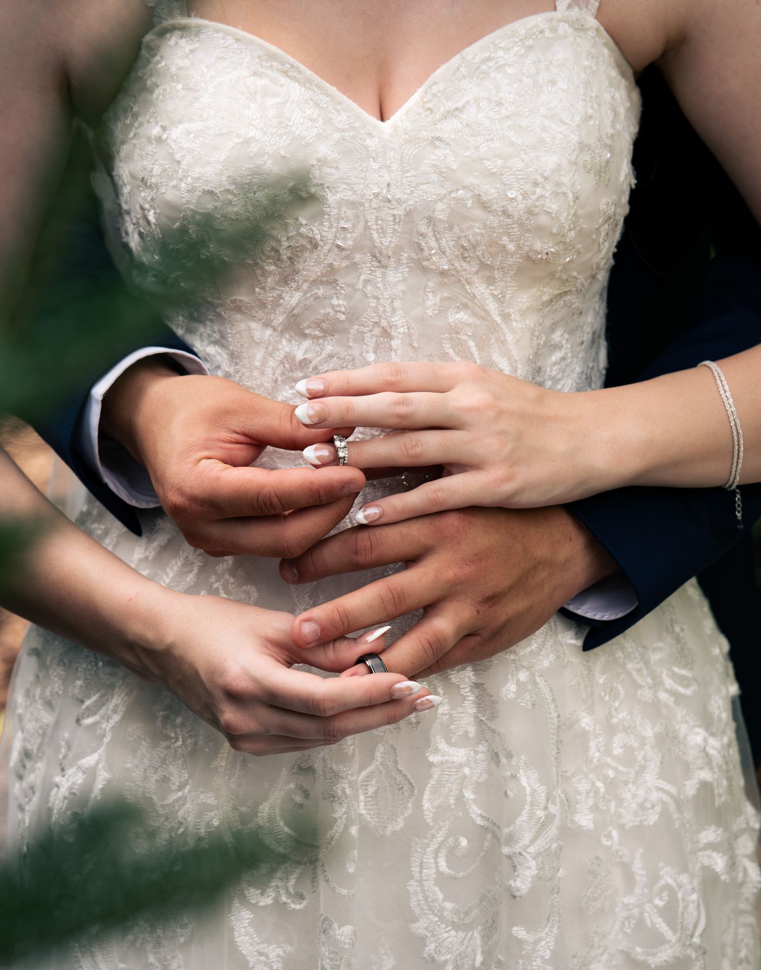 Bride and groom holding hands showing wedding rings, bride in lace gown, groom in navy suit.