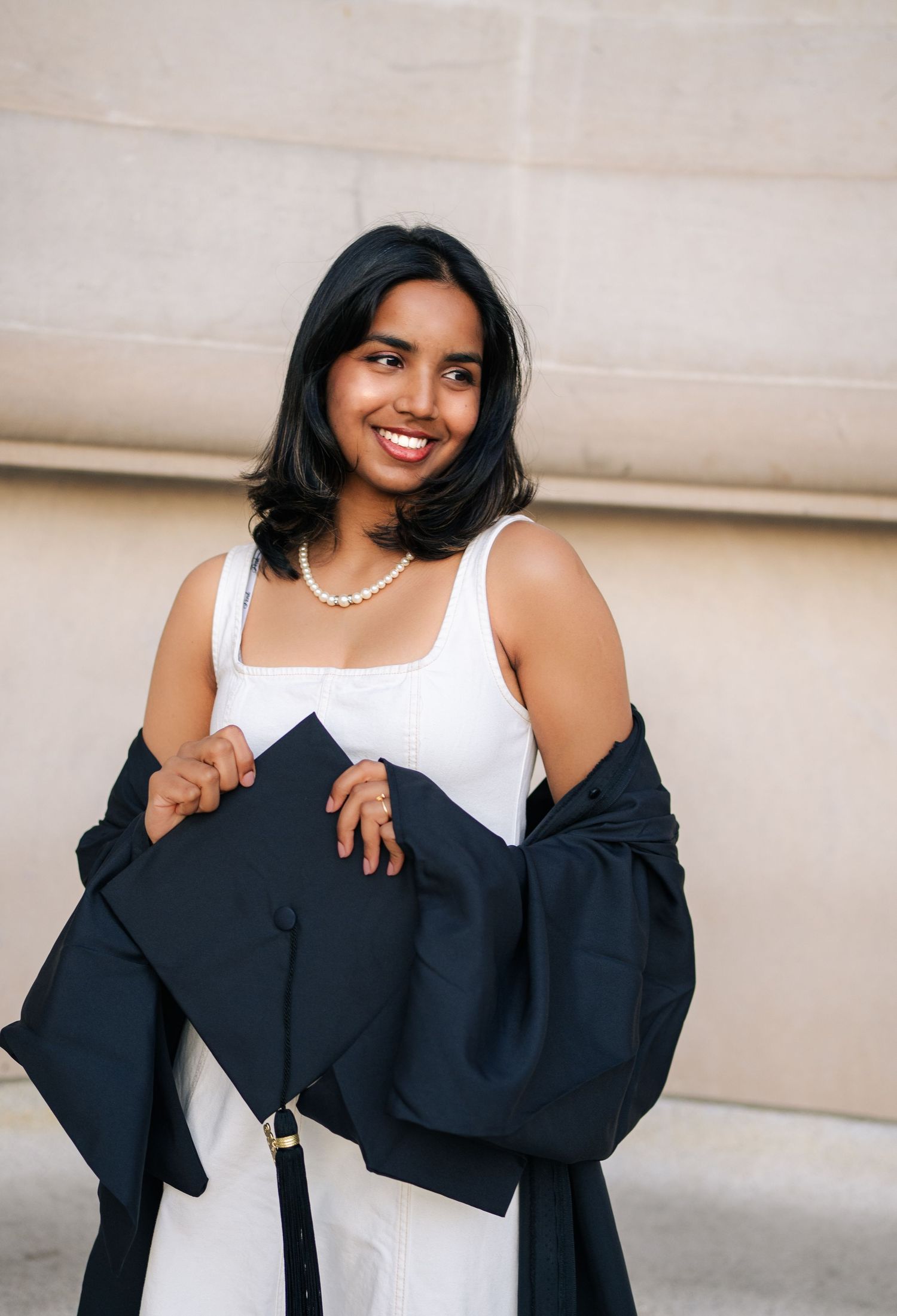 Smiling graduate in white dress and pearl necklace holds black cap and gown outdoors near stone wall.