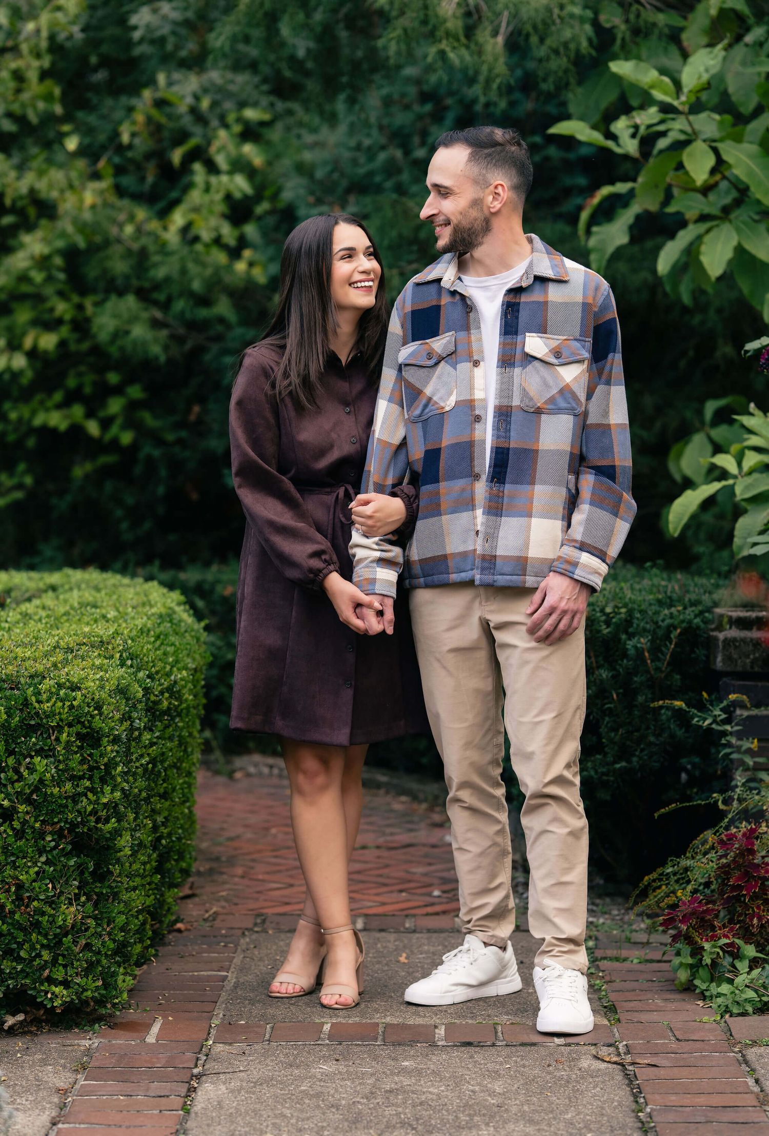 A couple smiles at each other while holding hands on a brick path surrounded by lush green gardens.