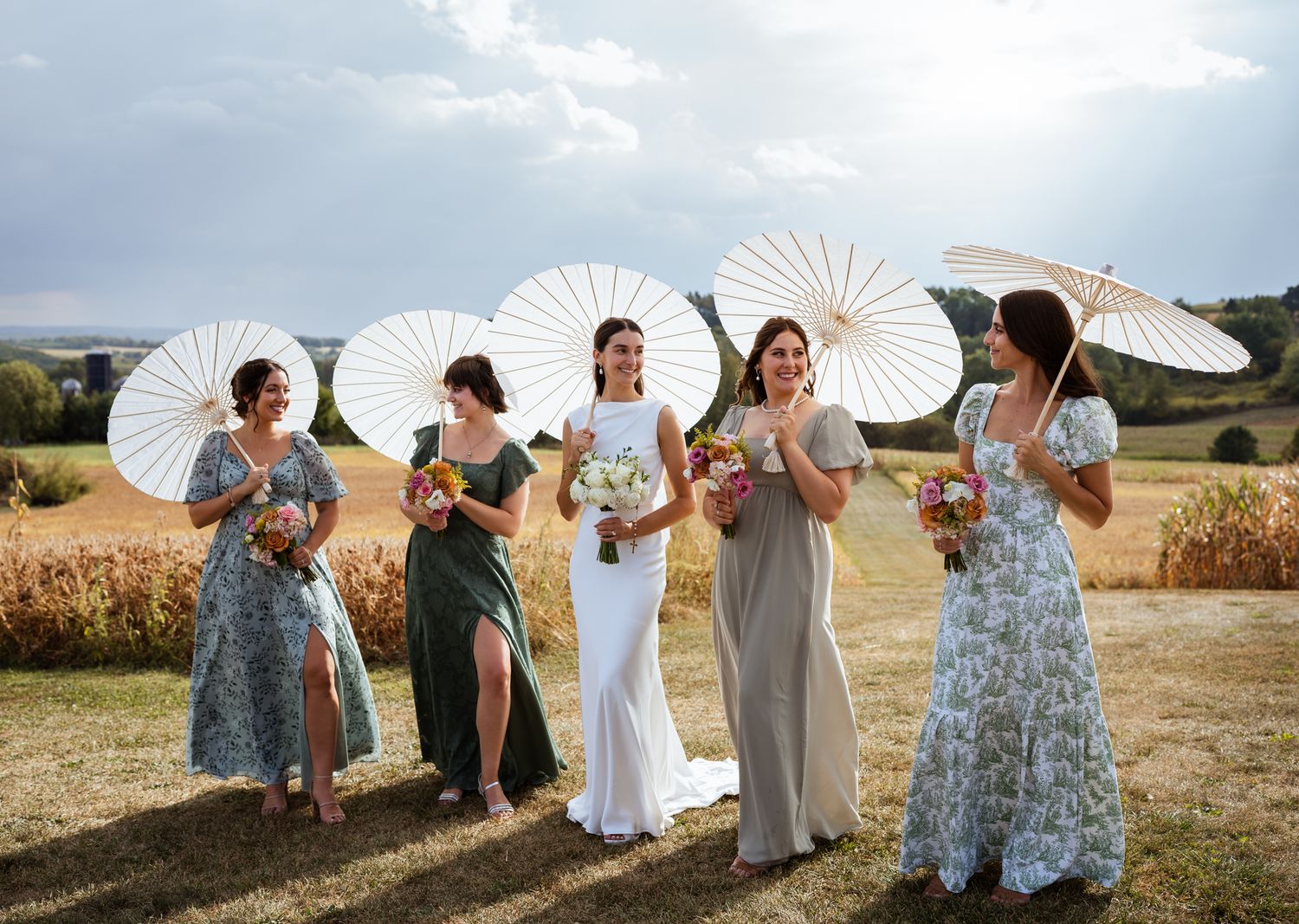 Bride and bridesmaids in floral dresses holding white parasols in a sunny countryside field.